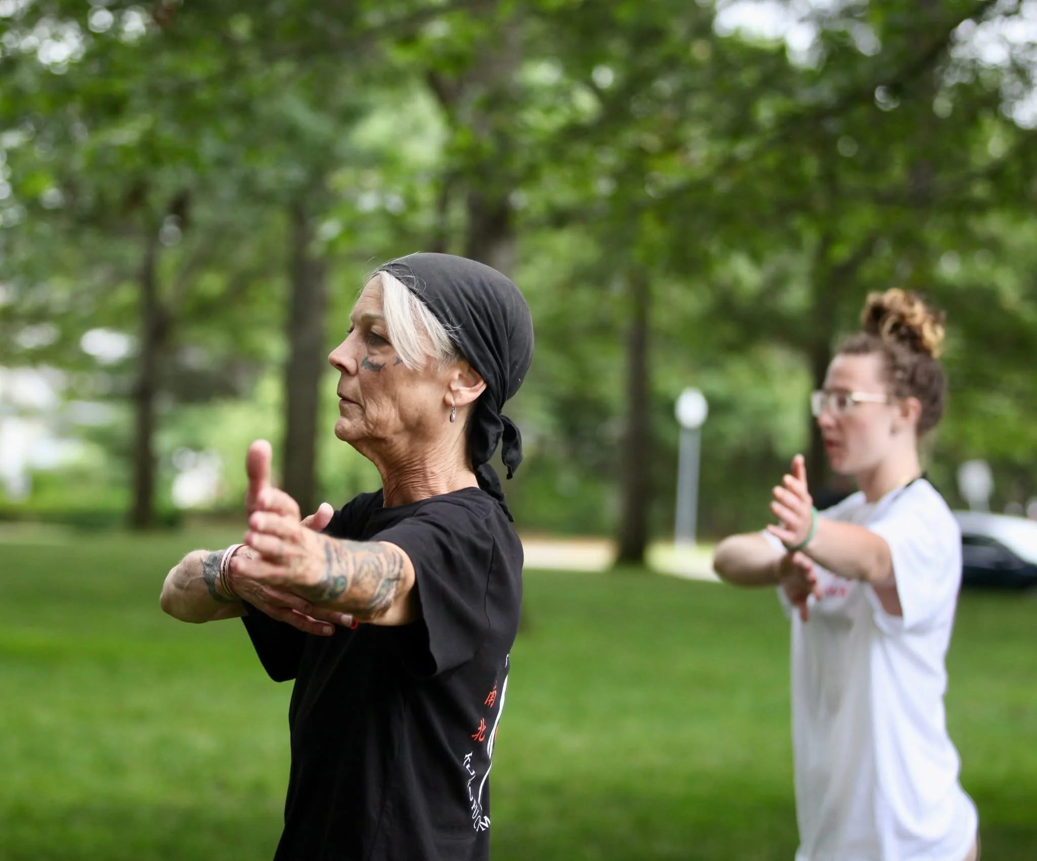 Two women practicing Tai Chi in a park during daytime, one older with gray hair wearing a black bandana, and one younger with glasses and a bun, both focused and in meditative poses.