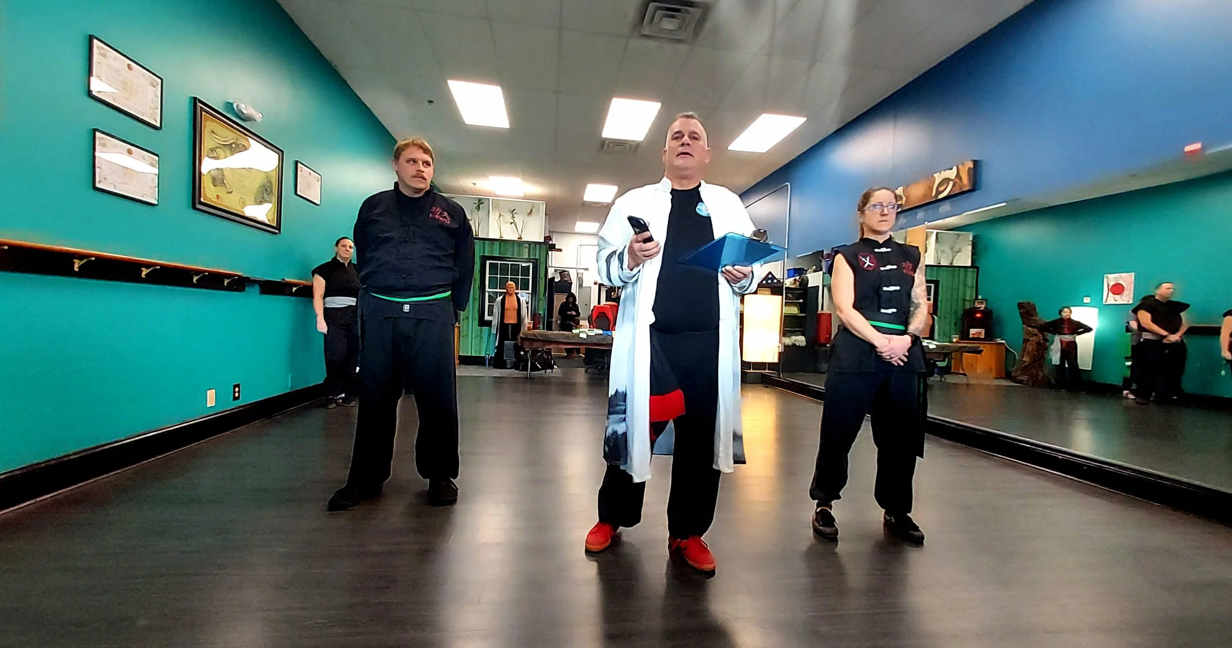 Group of people, including a man in a white coat holding a clipboard and a pen and a smartphone, standing in a room with teal walls, framed art, and a black floor, possibly a martial arts class or training session.