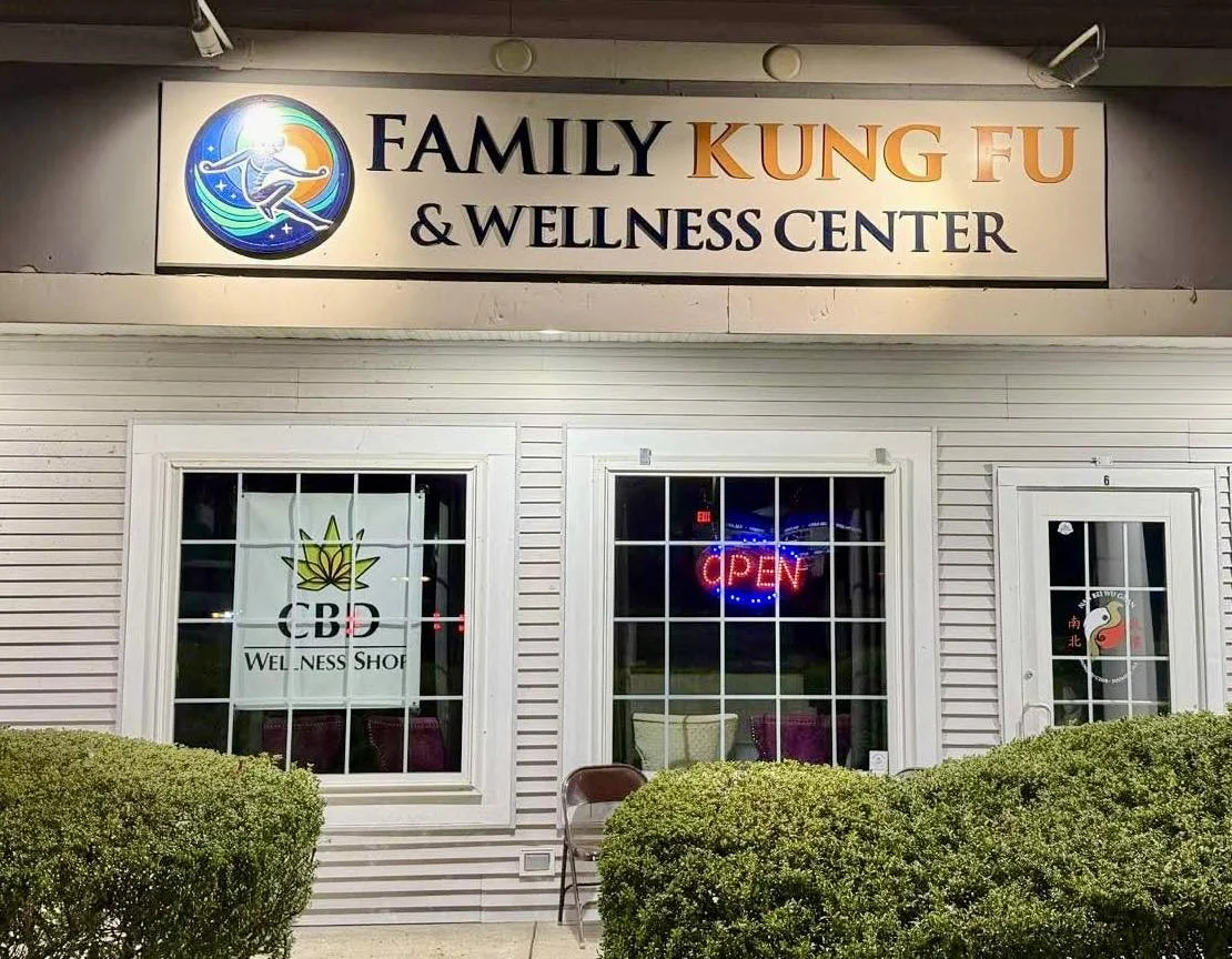 Exterior view of the Family Kung Fu & Wellness Center with a sign, windows, and a door. The center has an open neon sign and a cannabis wellness shop window.