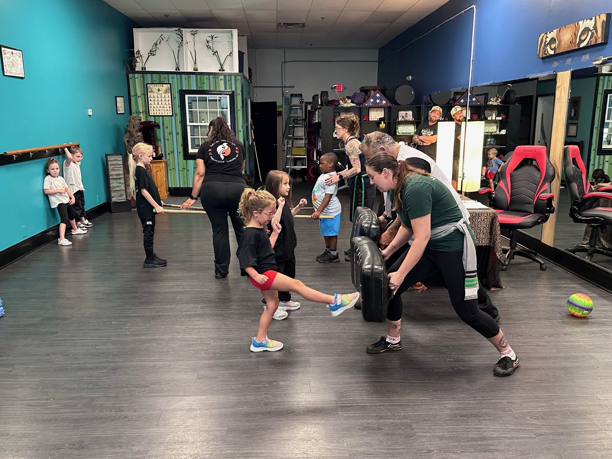 Children and instructors participating in martial arts class, practicing kicking techniques in a dance studio with blue and green walls, dark flooring, and martial arts equipment.