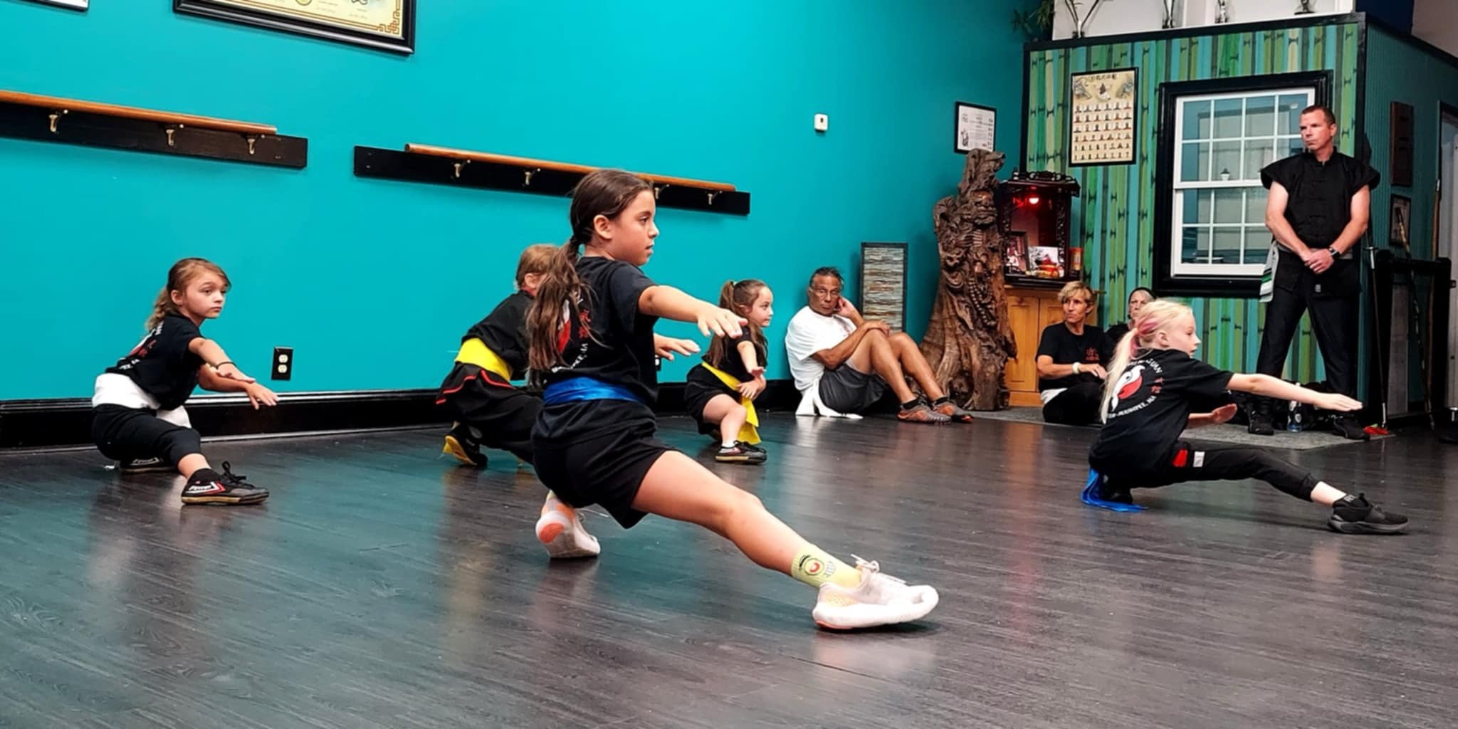 Children performing martial arts moves in a martial arts class with an instructor observing in a room with turquoise walls and wooden flooring.