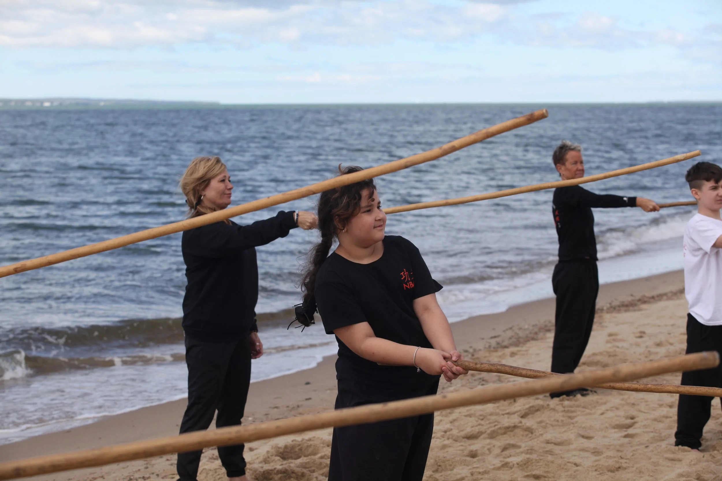 Group of four people practicing martial arts with wooden sticks on a sandy beach near the water, under a cloudy sky.