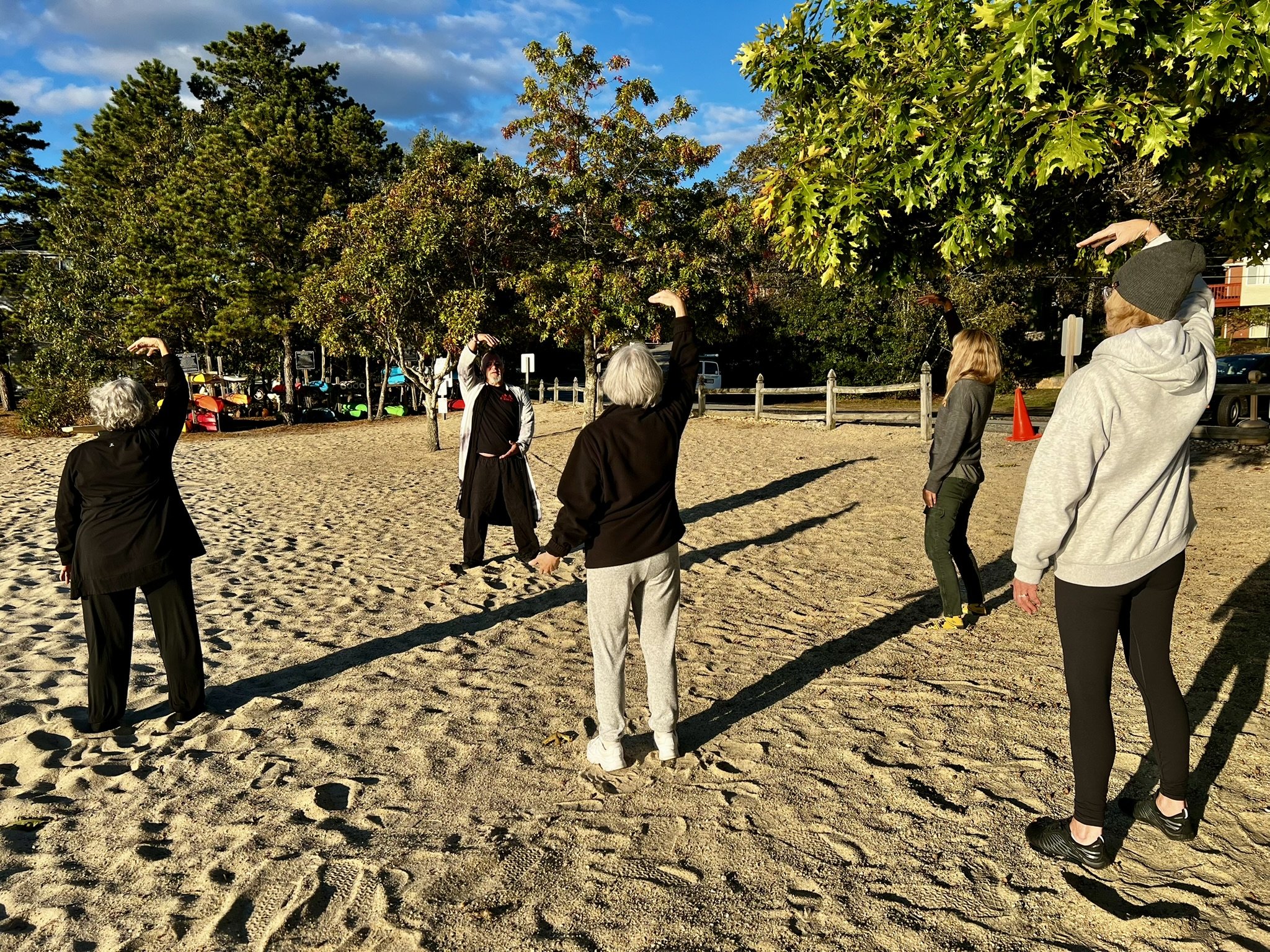 Group of six people practicing Tai Chi or stretching exercises on a sandy outdoor area with trees and parked cars in the background during late afternoon.