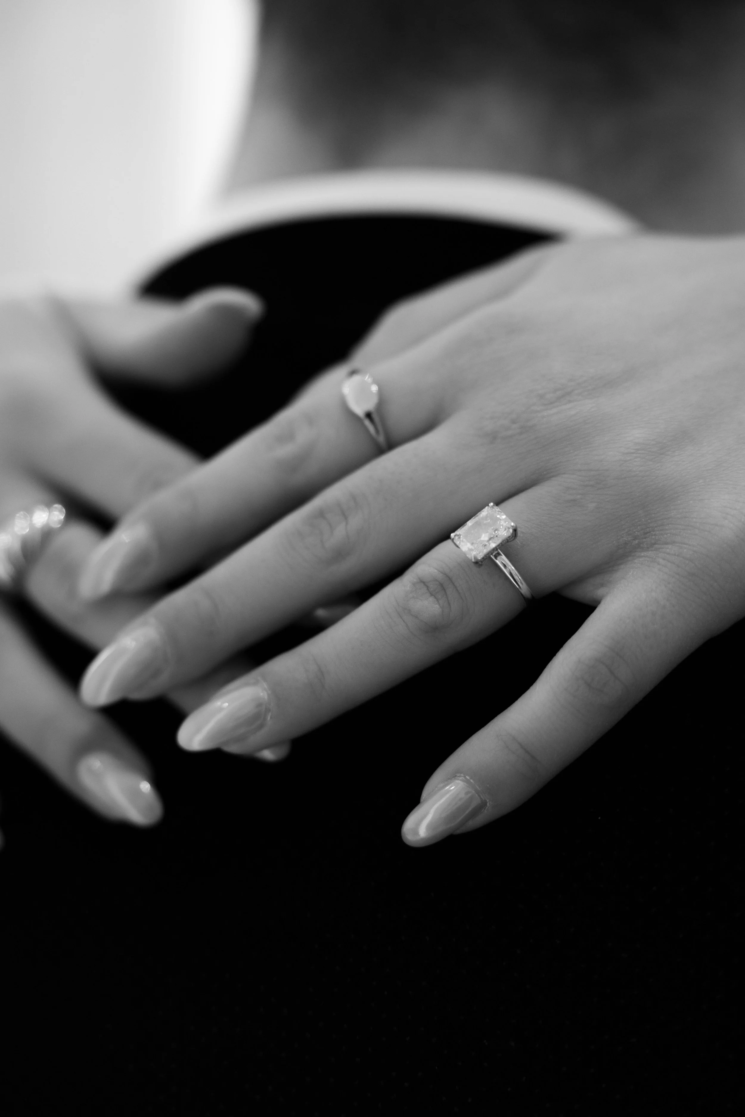 Close-up of hands with rings, one with a large rectangular gemstone, on a dark surface in black and white.
