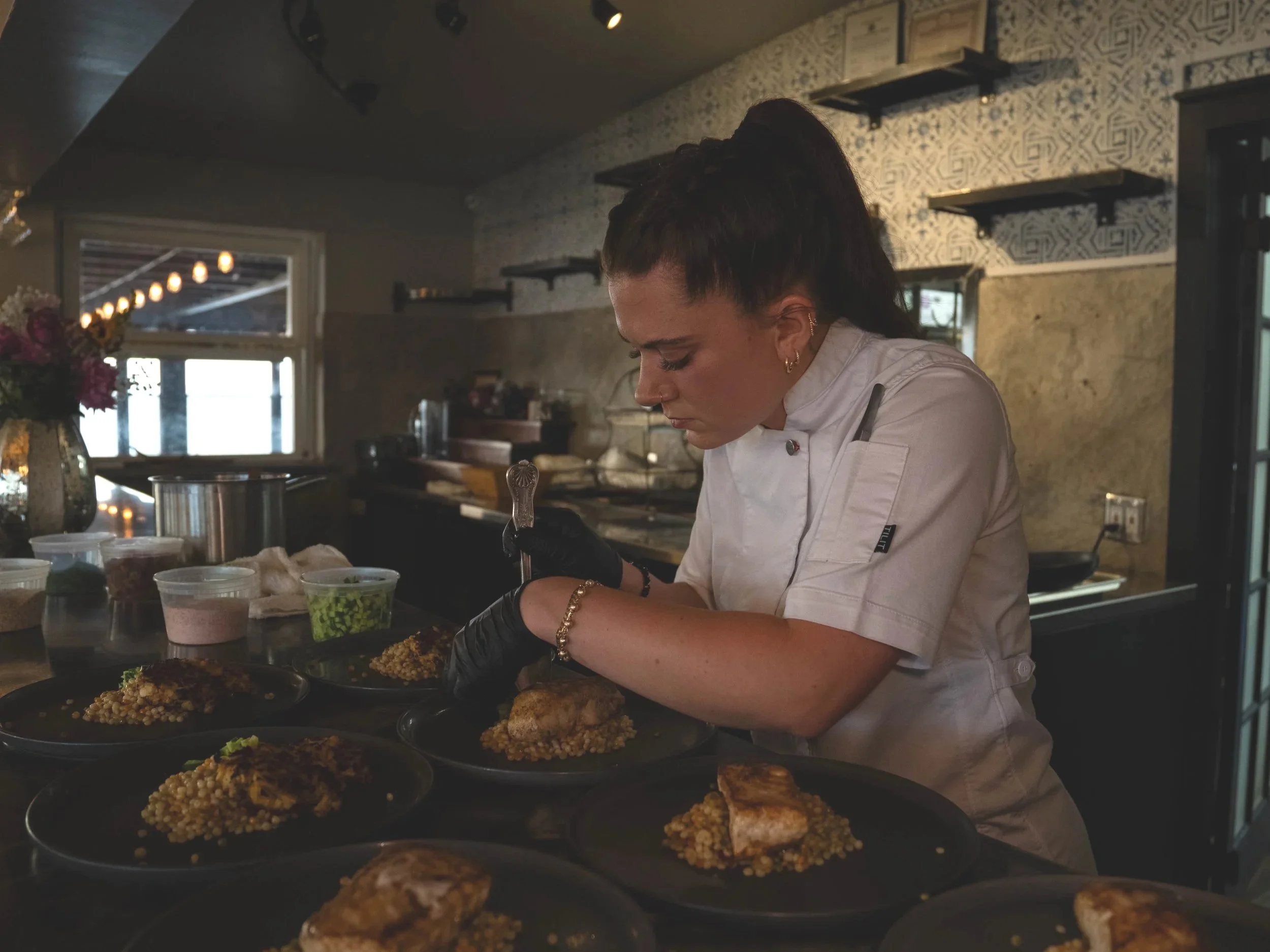 A chef wearing a white uniform and black gloves is plating food in a restaurant kitchen with plates of couscous and chicken.