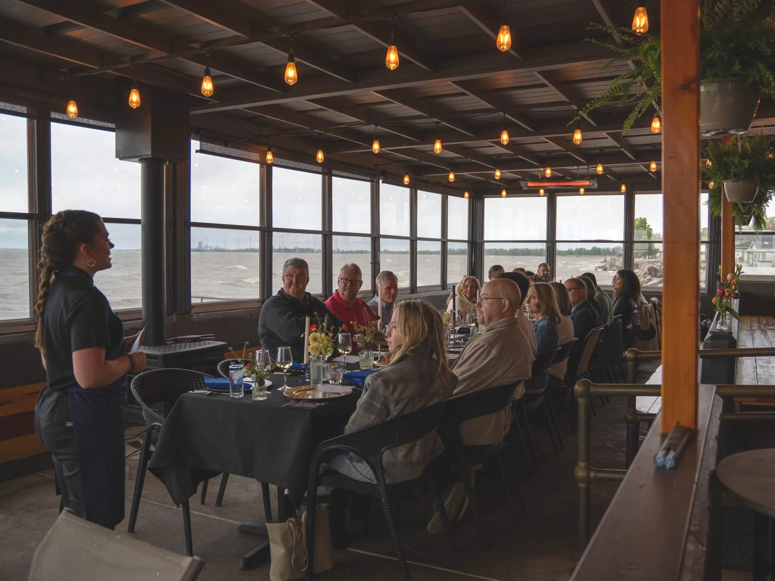 Group of people attending a meeting or dinner in a restaurant with large windows showing a body of water and city skyline in the background, and a woman standing and speaking to the group.