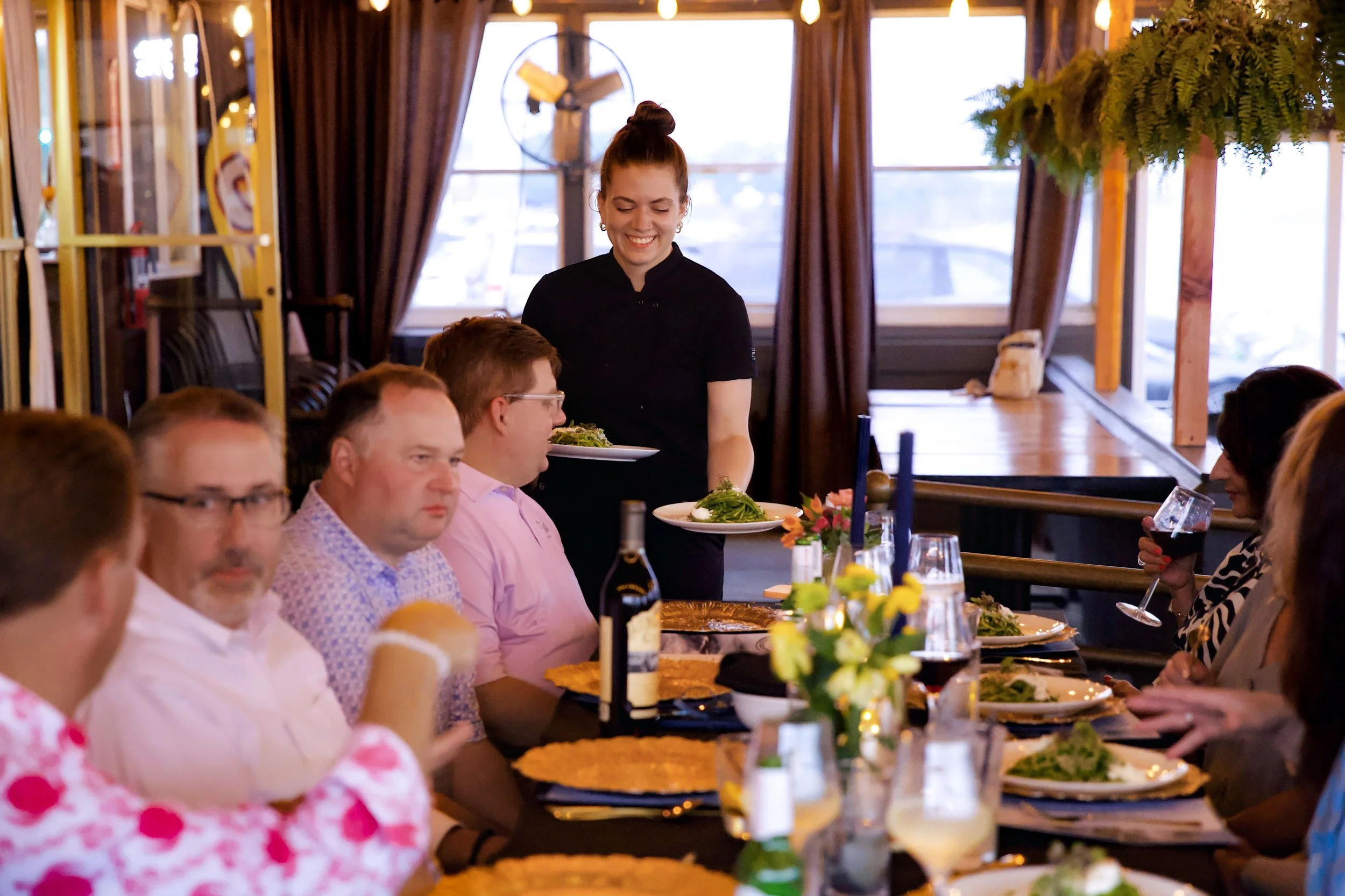 A woman in black serving food at a restaurant table to diverse people, with a table set with plates of salad, drinks, and a decorative centerpiece.