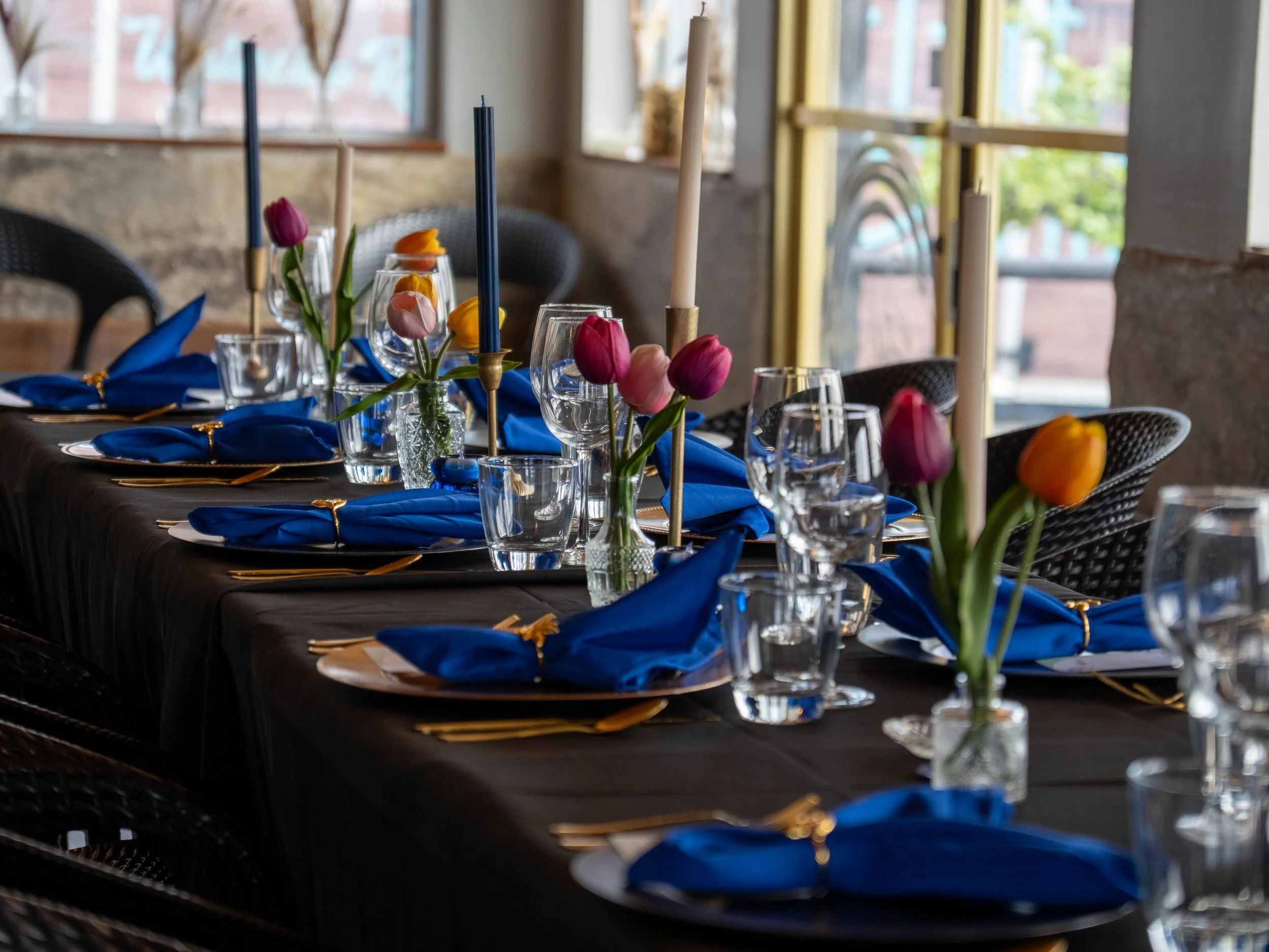 A long dining table decorated with blue napkins, glassware, colorful tulips in vases, and tall candles, set up for a formal event in a room with large windows.