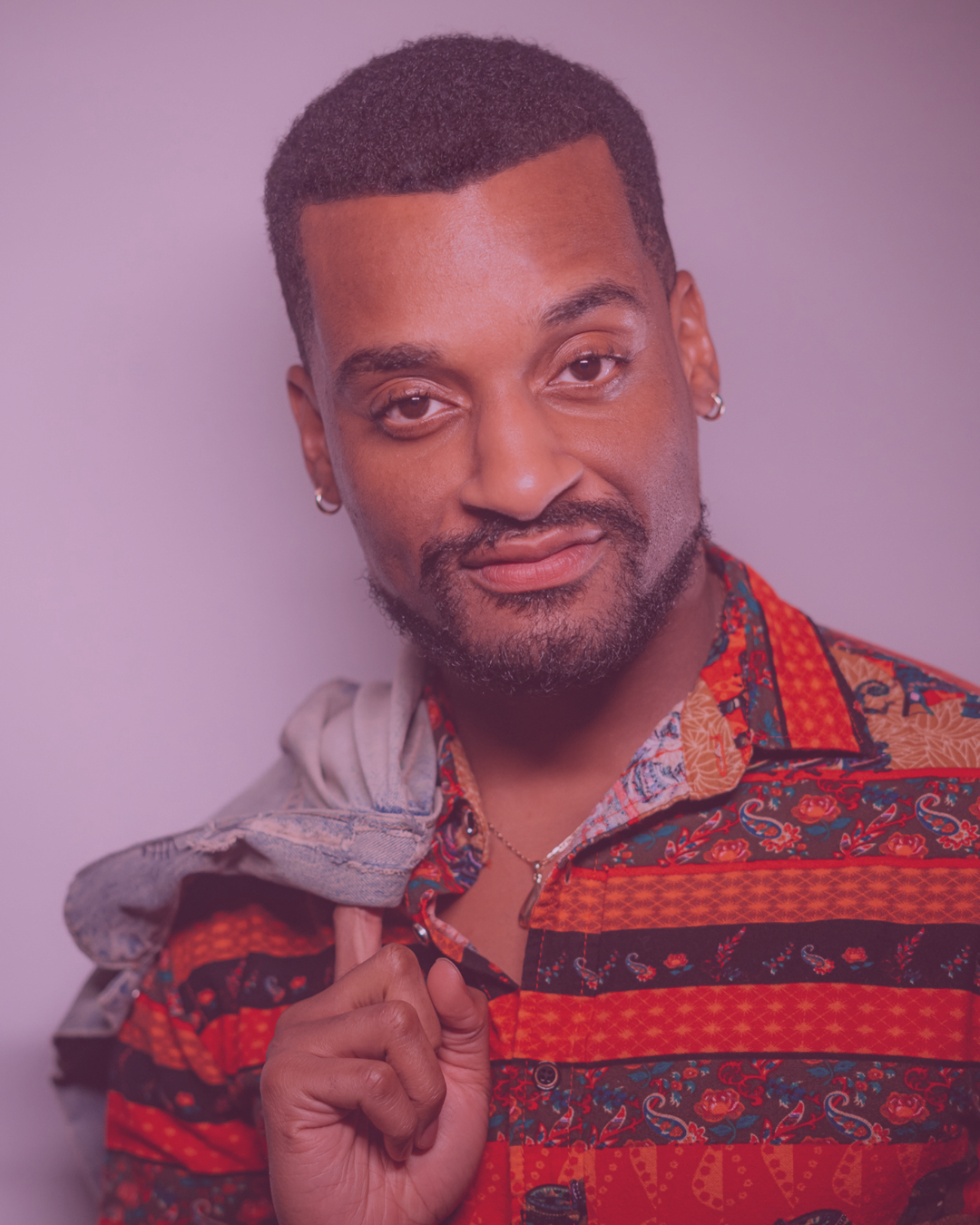 Ace Anderson. A man with short hair and earrings, wearing a colorful patterned shirt, holding a denim jacket over his shoulder, smiles at the camera.