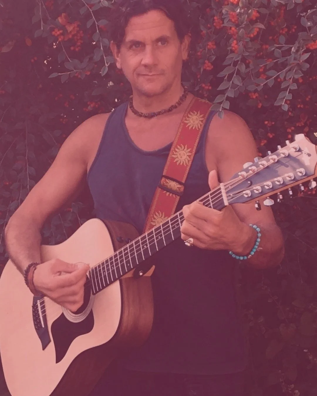 A man wearing a sleeveless gray shirt, holding an acoustic guitar, standing in front of a bush with red berries. He has a beaded bracelet on his right wrist, a beaded necklace, and a colorful guitar strap with a sun design.