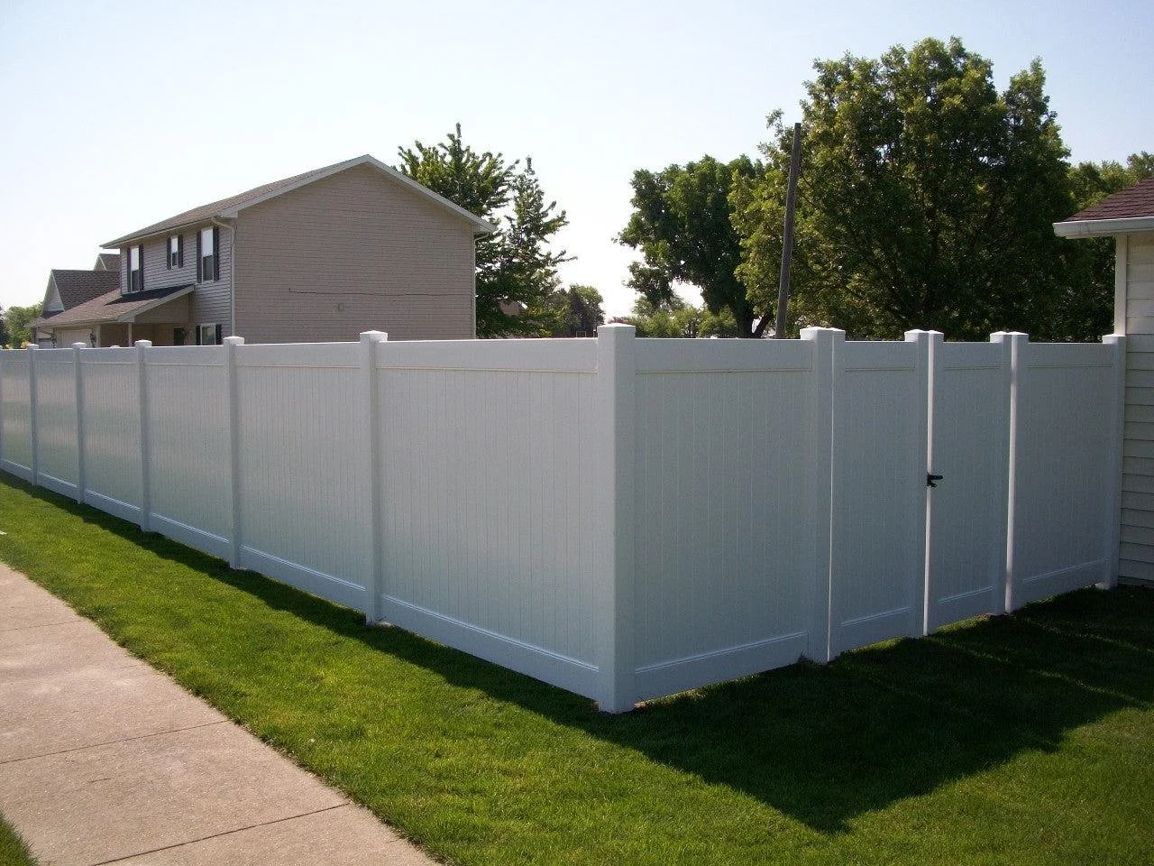 White vinyl privacy fence along a sidewalk next to a house with siding, with green grass, trees, and a clear sky in the background.