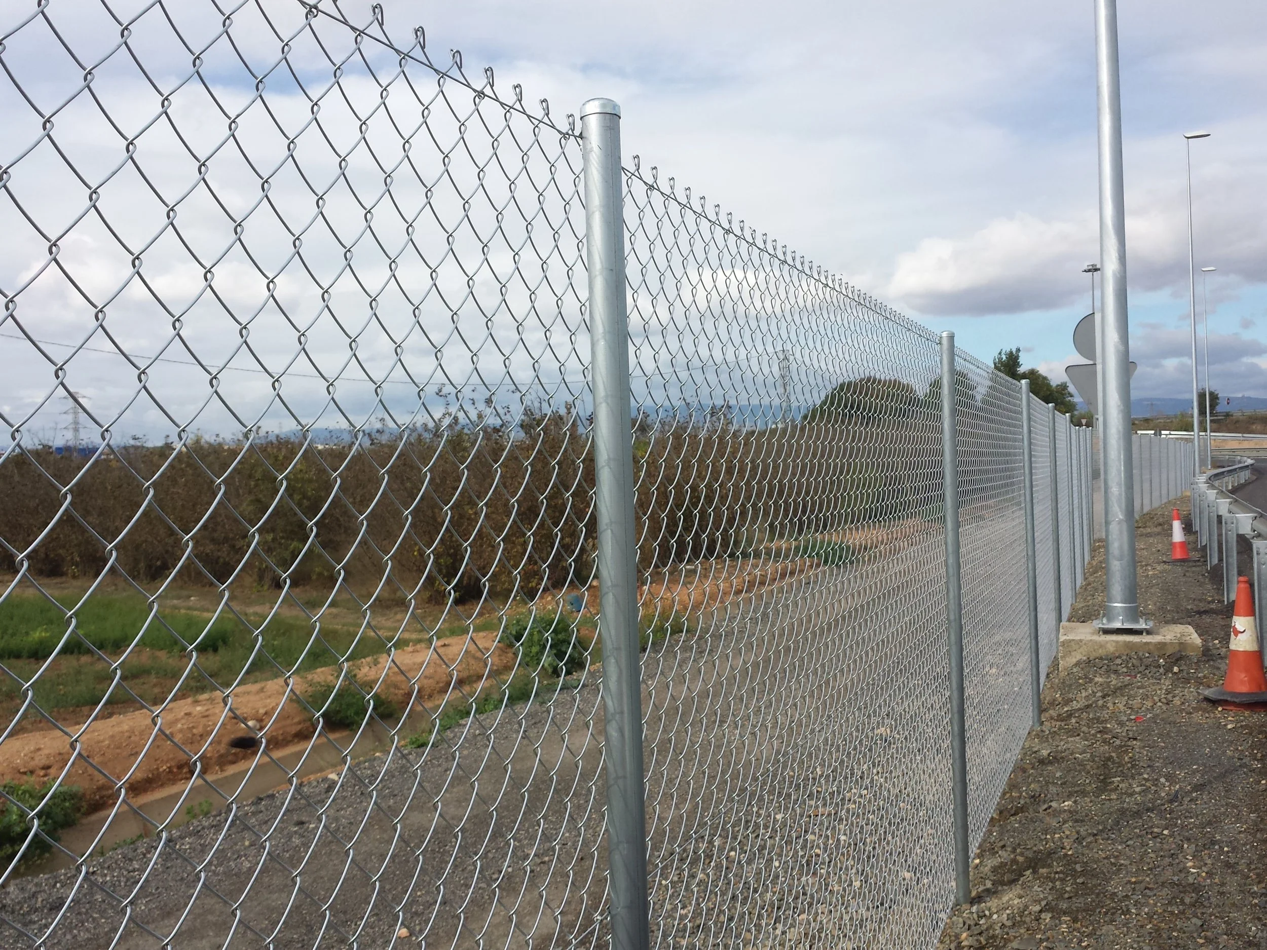 Metal chain-link fence along a construction or road area with orange traffic cones, open land with bushes and trees, cloudy sky in the background.