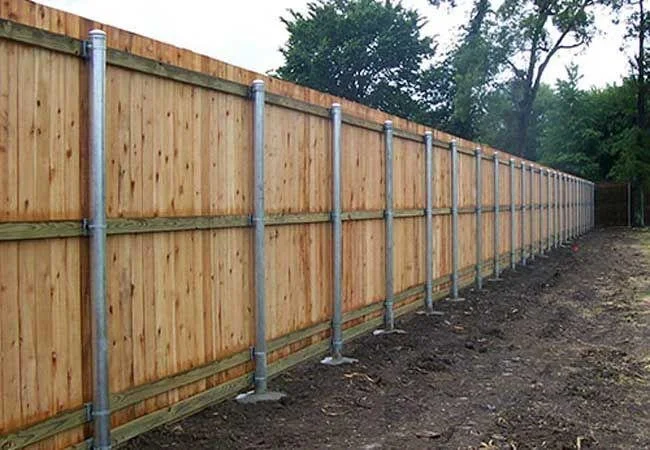 Newly installed wooden privacy fence with metal support posts along a dirt ground and trees in the background.