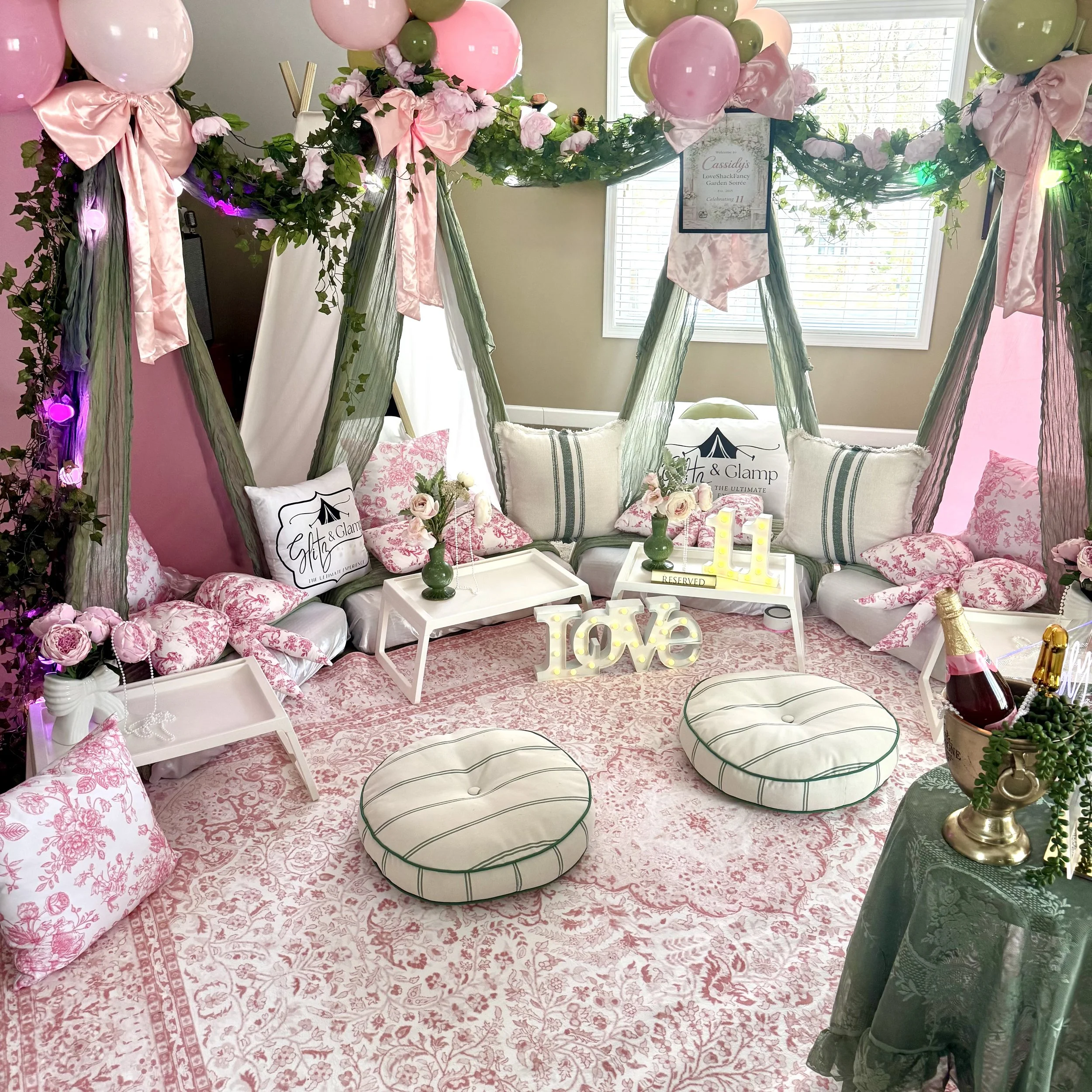 Decorative setup with pastel pink and green balloons, large pink ribbons, floral accents, and reserved sign on a small white tray in front of three bed-like arrangements, in a room with green walls and dark furniture.
