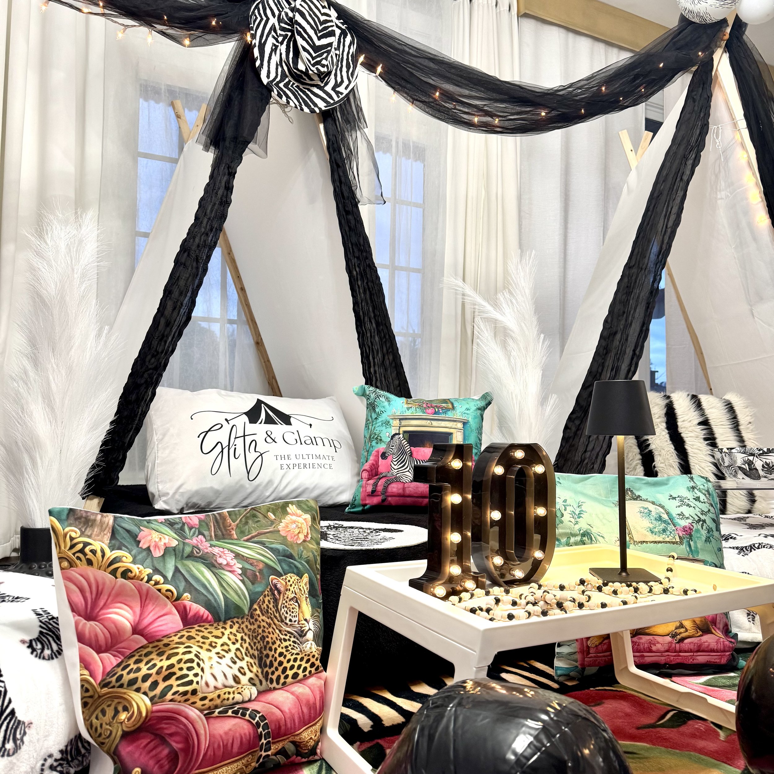 Decorative interior setting featuring a black and white tent-like structure with black mesh fabric and string lights, colorful pillows with animal and nature themes, a white table with a lit marquee '10' number, a black table lamp, and dried white pampas grass.