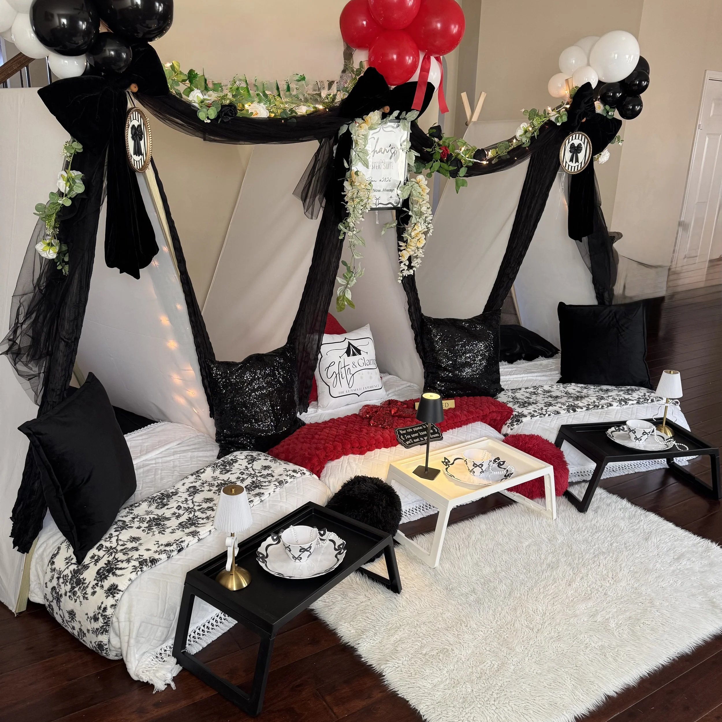 Decorative seating area with cushions and small tables surrounded by black and white balloons, sheer fabric, and floral arrangements.