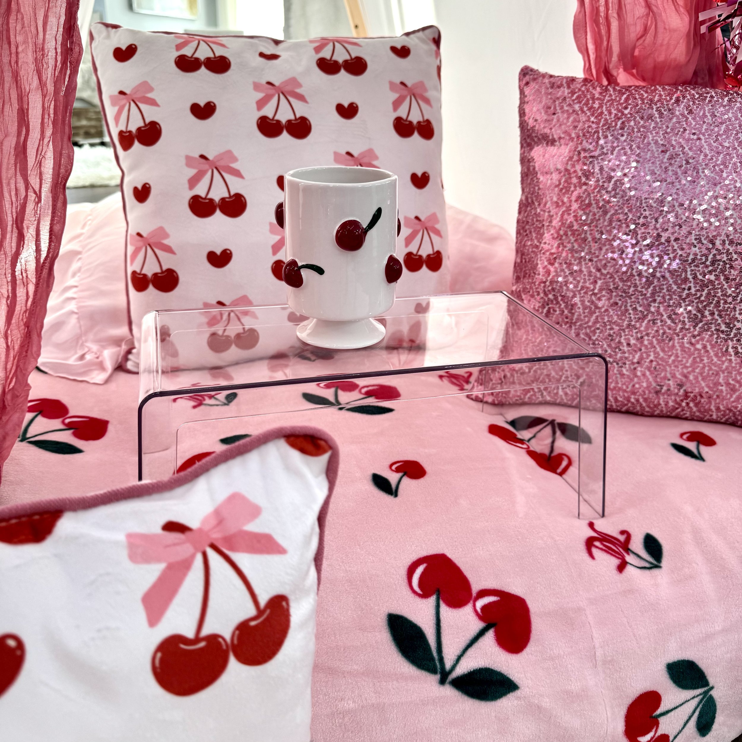 A bedroom with cherry-themed decor, featuring pink and white bedding with cherry print, matching pillows, and a clear acrylic table displaying a white cup decorated with cherries.