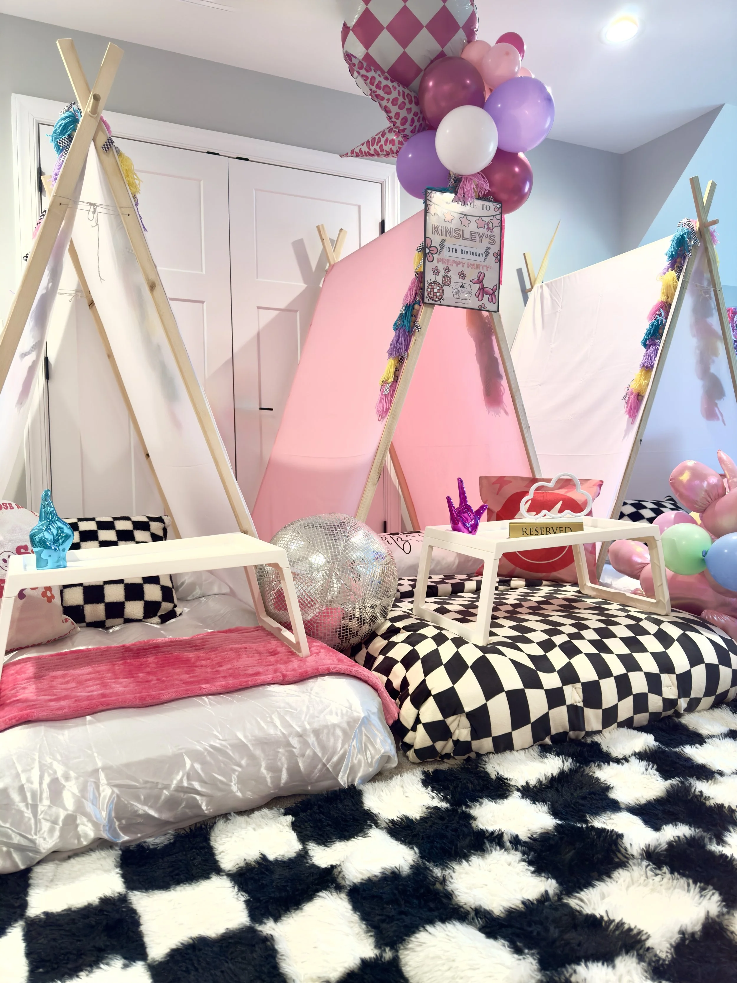 Decorative indoor scene with three white teepees adorned with colorful pink and orange balloons, neon heart signs, and smiley face pillows, set on a fluffy white rug.