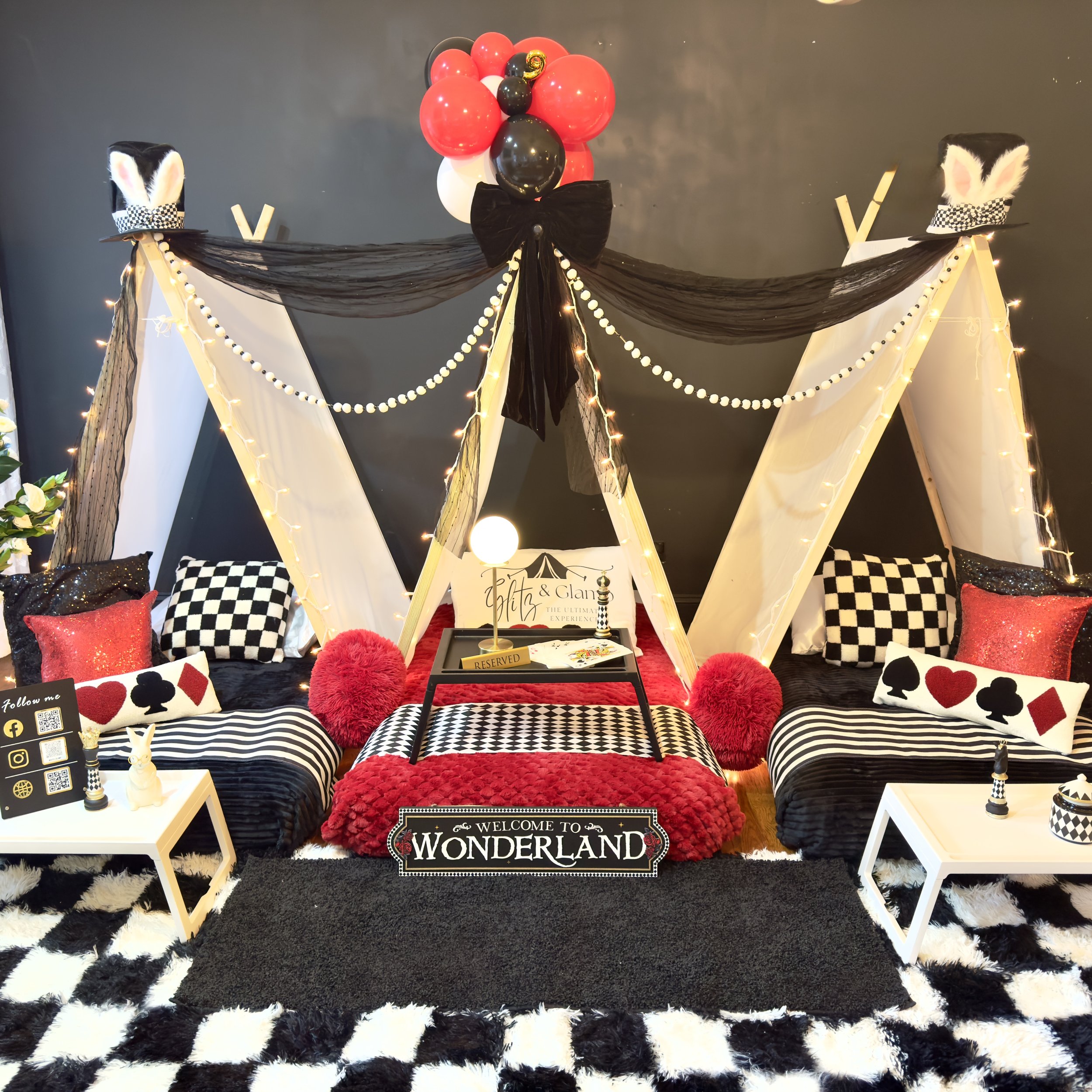 A whimsical, Alice in Wonderland-themed indoor setup with black and white tents, red and black balloons, and decorative pillows featuring hearts, clubs, spades, and diamonds, alongside fairy lights and playful bunny ears on top hats.
