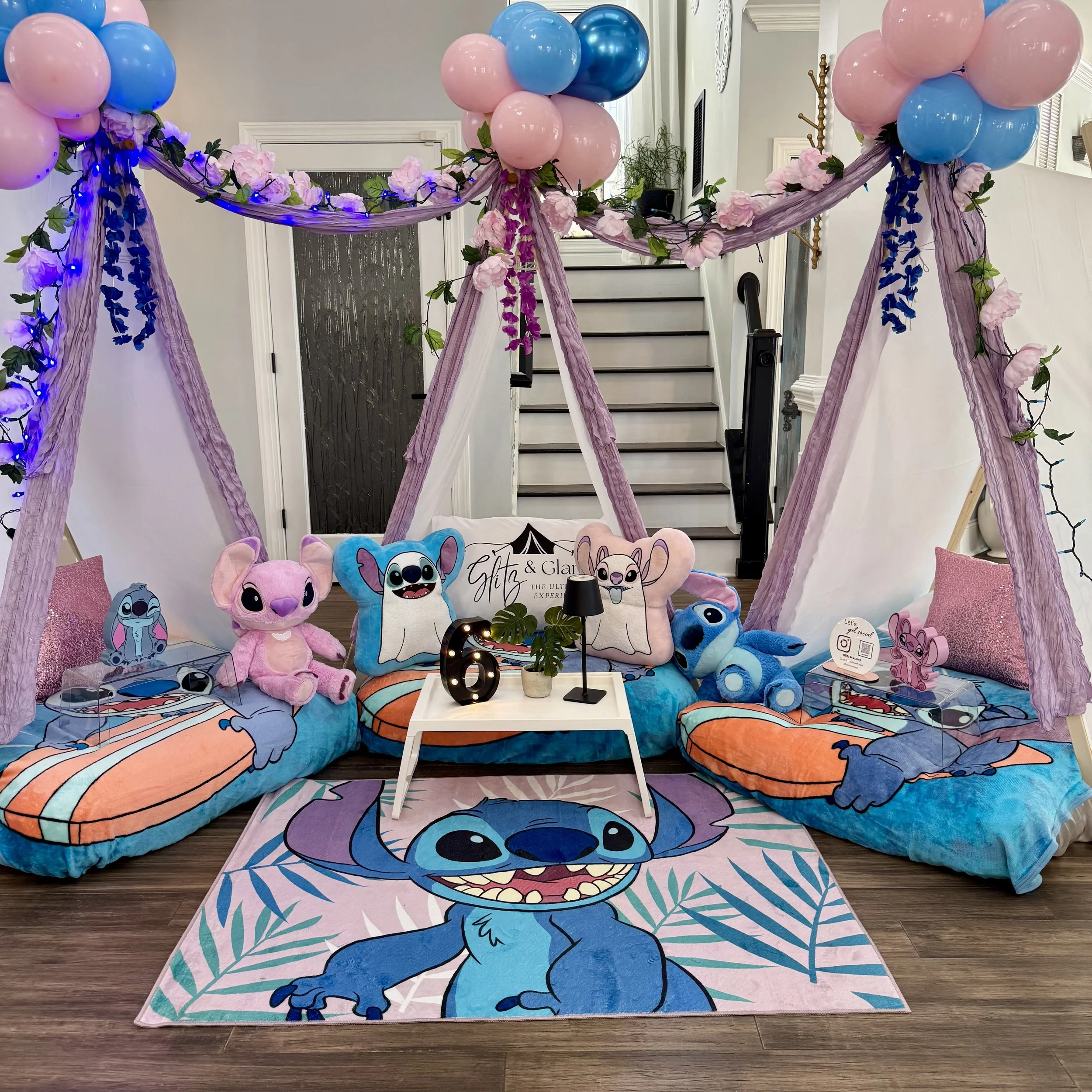 Decorative setup with Stitch from Lilo & Stitch theme, featuring plush toys, pillows, and a rug with Stitch's face. Pink, blue, and purple balloons and floral garlands form a canopy. Small tables with decorative items are on either side. The setting appears to be hosted indoors with stairs in the background.