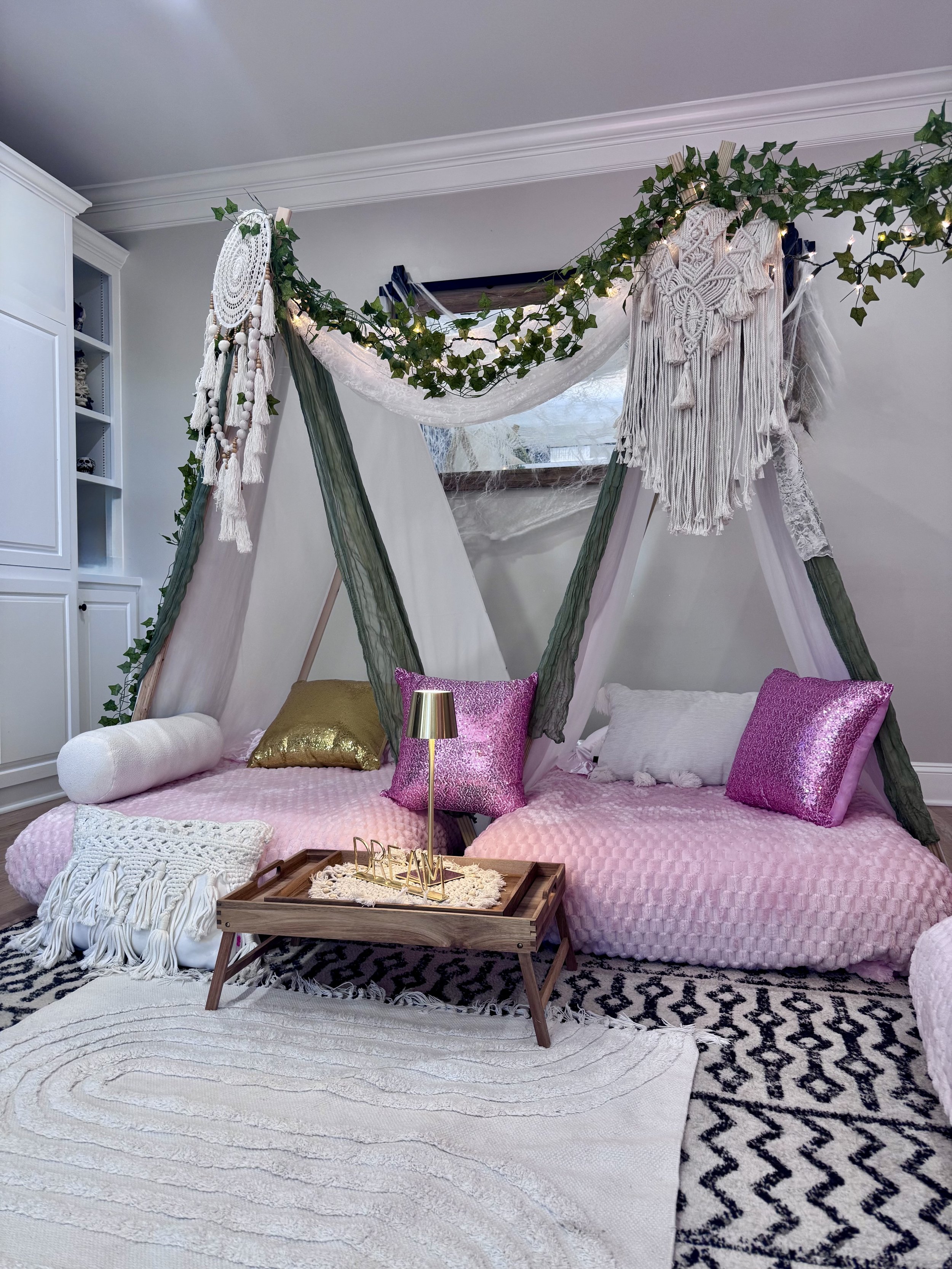 Indoor bohemian-style canopy tent with pink cushions, gold and pink pillows, white and gold throws, and decorative canopy with greenery, macrame, and string lights, over a low wooden table with a lamp and 'DREAM' decor, on patterned rugs, in a white room.