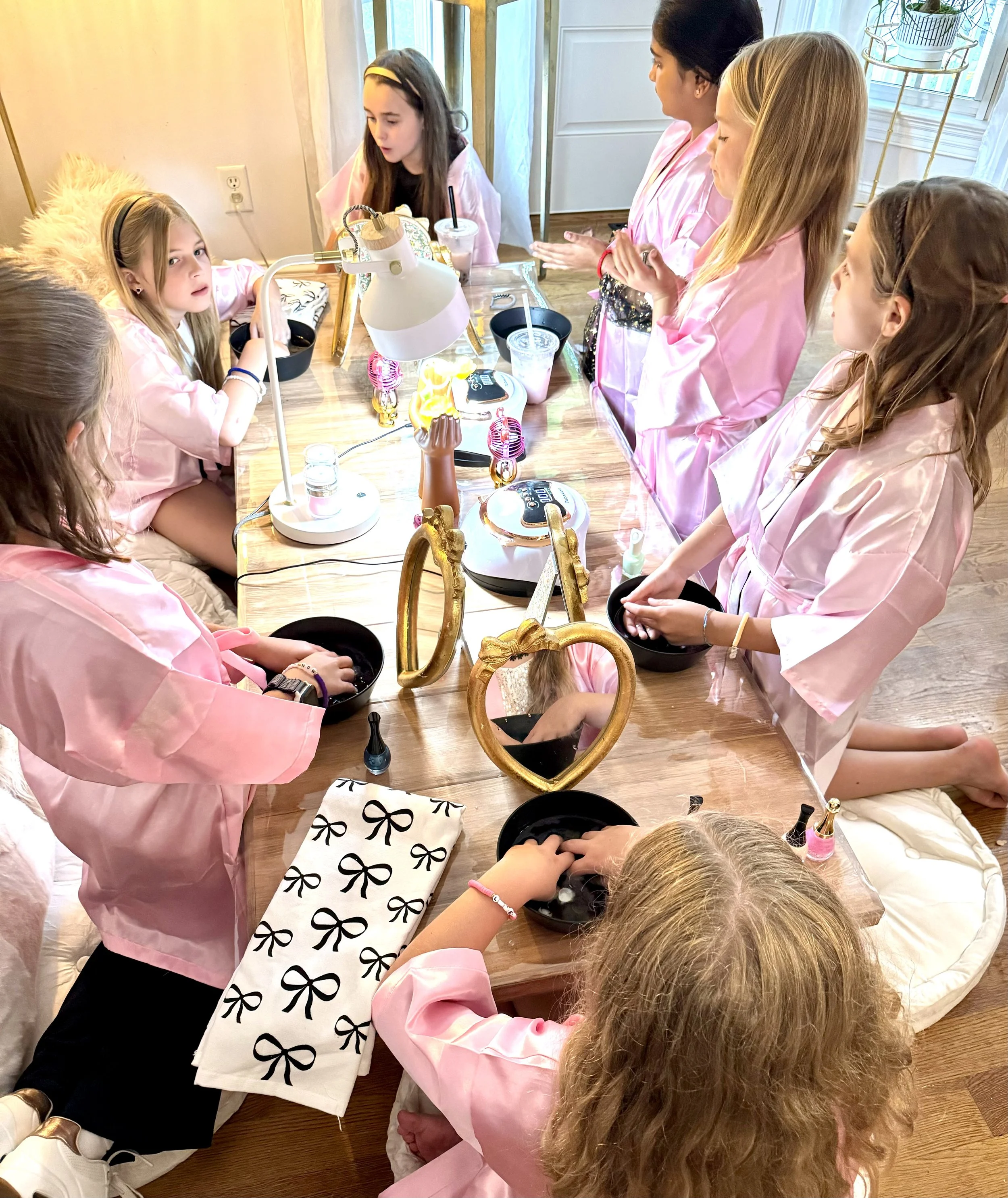 Four young girls in pink robes and purple headbands sitting together during a celebration with pink balloons and pink tissue paper pom-poms in the background.