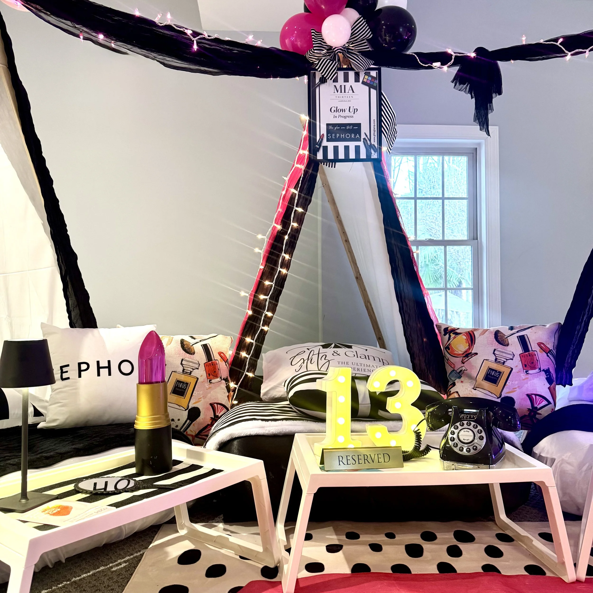 A birthday setup featuring two black and white striped teepees with big striped bows on the top, decorated with balloons in black, pink, and gold, along with pillows and a white rug with black text that reads 'SPA DAY EVERYDAY'.