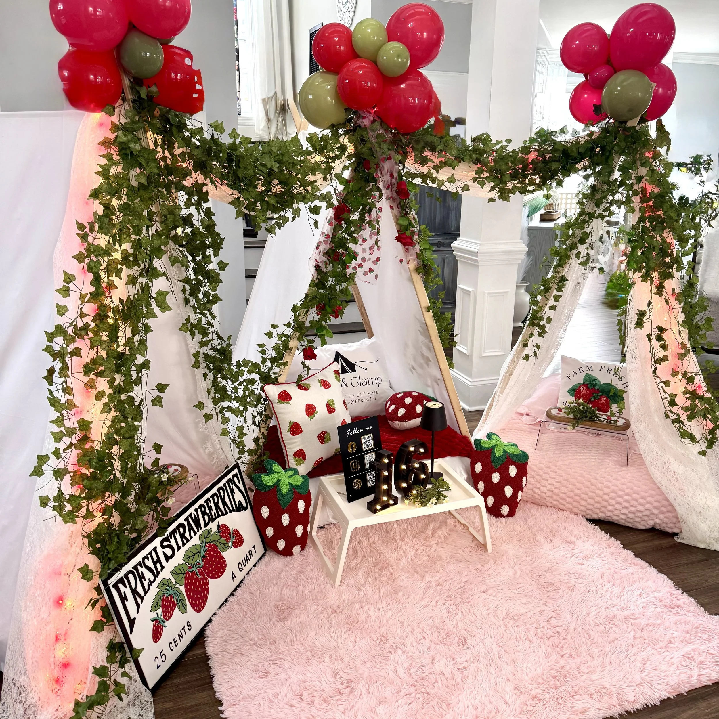 Decorative setup with strawberries, ivy, and pink elements, including a small tent, pillows, and signs.