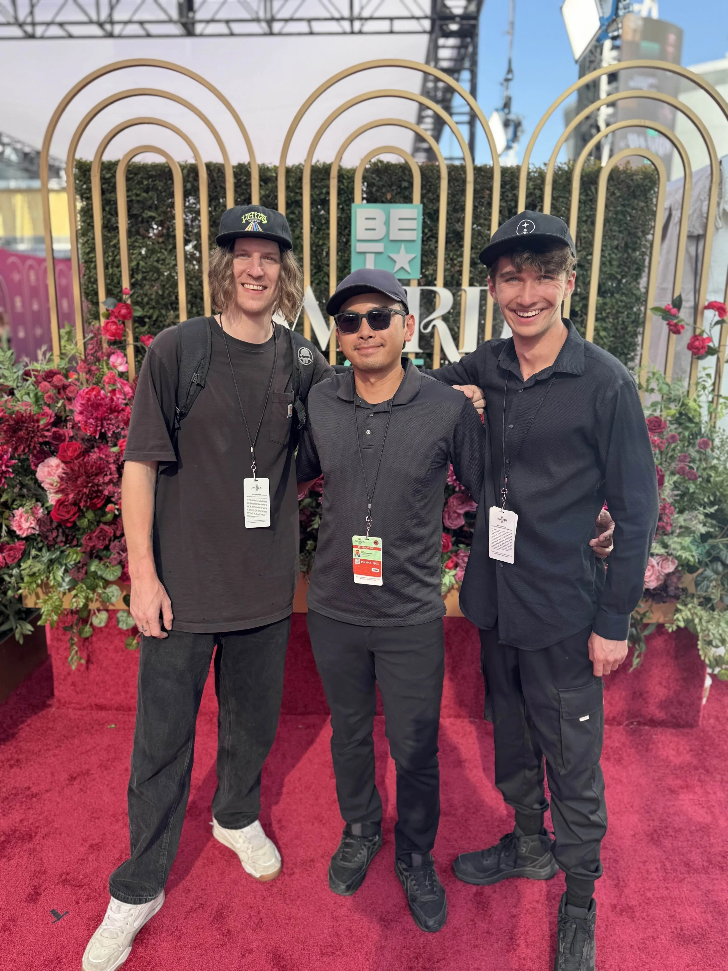 Three men standing together at a BET Experience event, with a backdrop of pink flowers, green foliage, and a sign that reads 'BET' and 'MORNING'. They are smiling, dressed in black, and wearing badges around their necks.