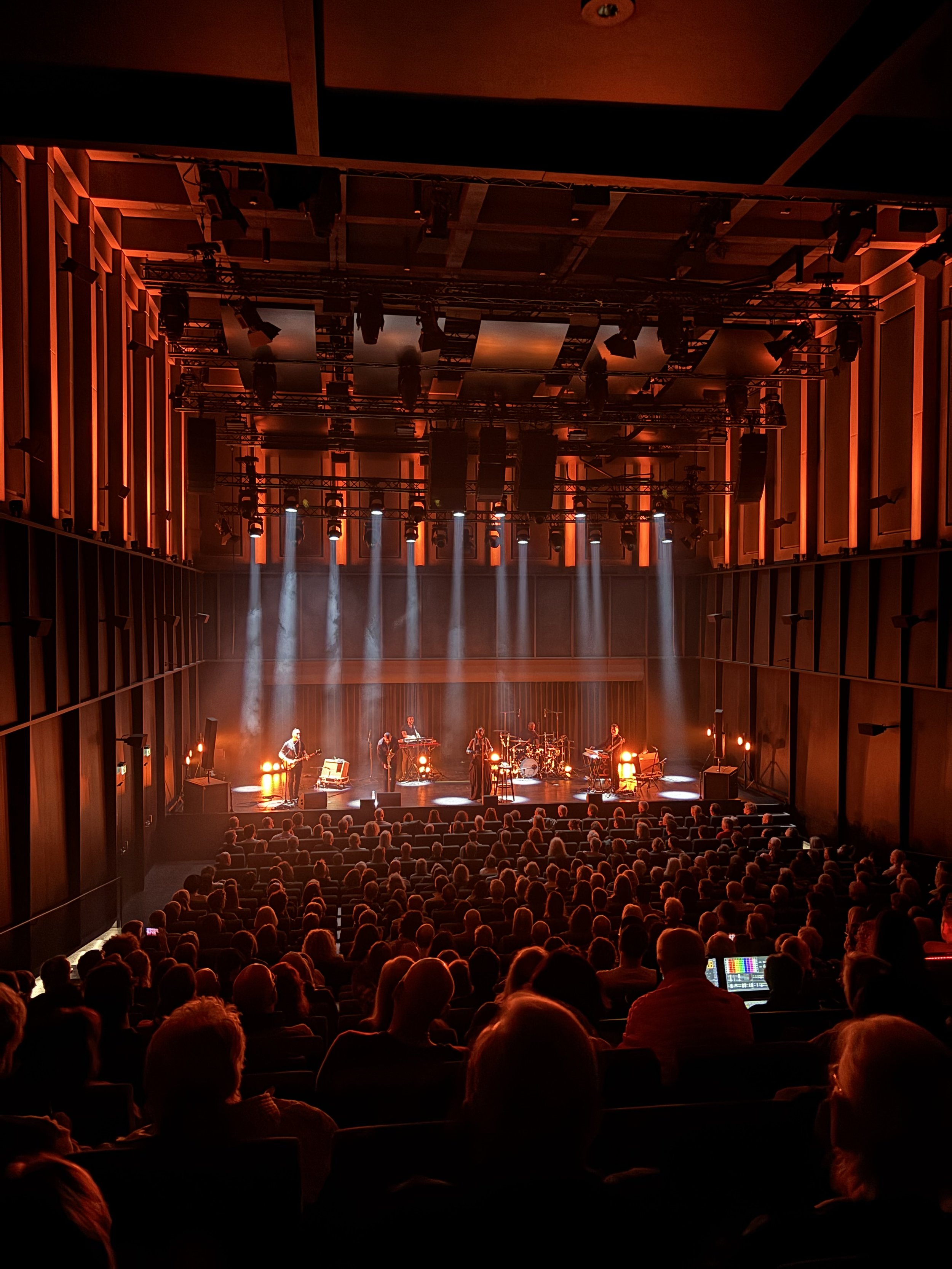 A live concert in a modern theater with a band performing on stage, dramatic lighting, and a large audience seated in rows facing the stage.
