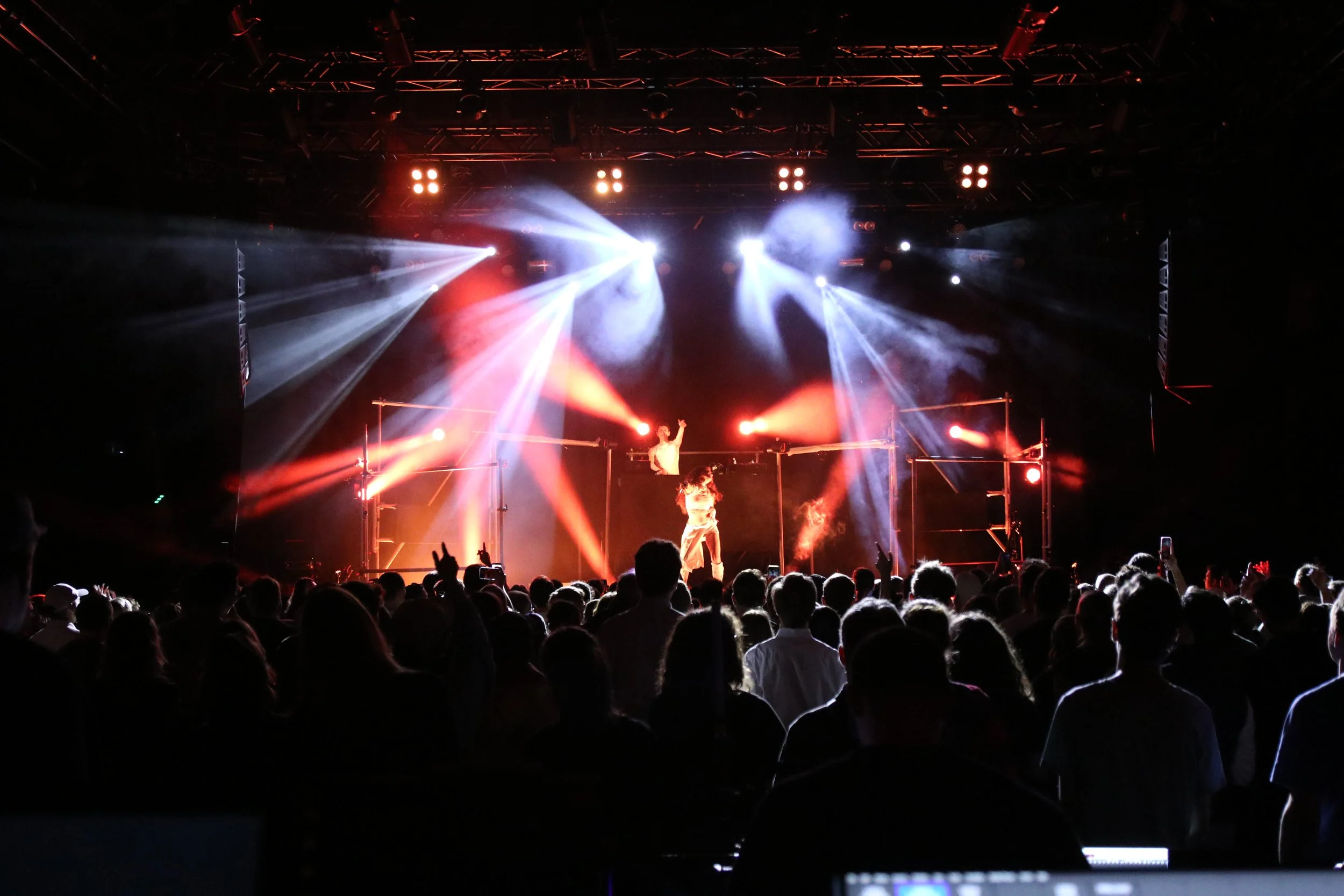A concert or music festival scene with a large crowd facing a stage illuminated with colorful, bright lights. There is a performer on stage, possibly a DJ or singer, with fog or smoke effects and various lighting effects creating a vibrant atmosphere.