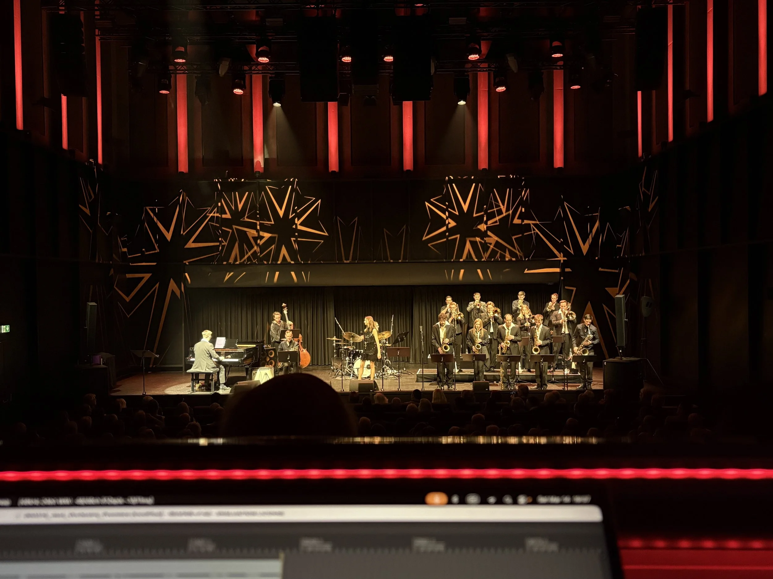 A large stage with musical performers, including a pianist, saxophonists, and a drummer, decorated with a black and gold star pattern, illuminated with red lighting and vertical red accents in the background, during a concert or performance.