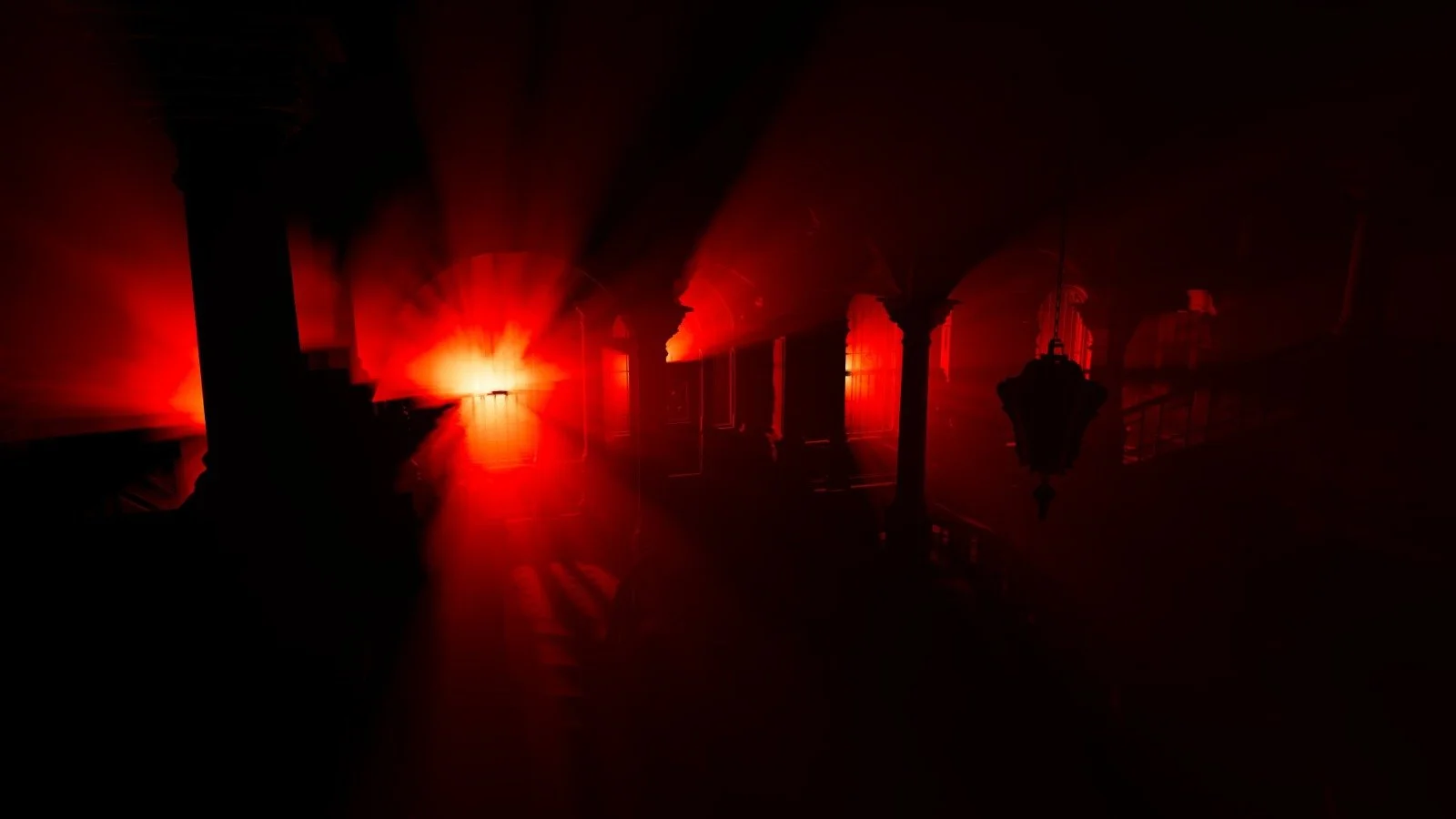Dark hallway illuminated by intense red light, with arched doorways and hanging lanterns, creating an ominous atmosphere.