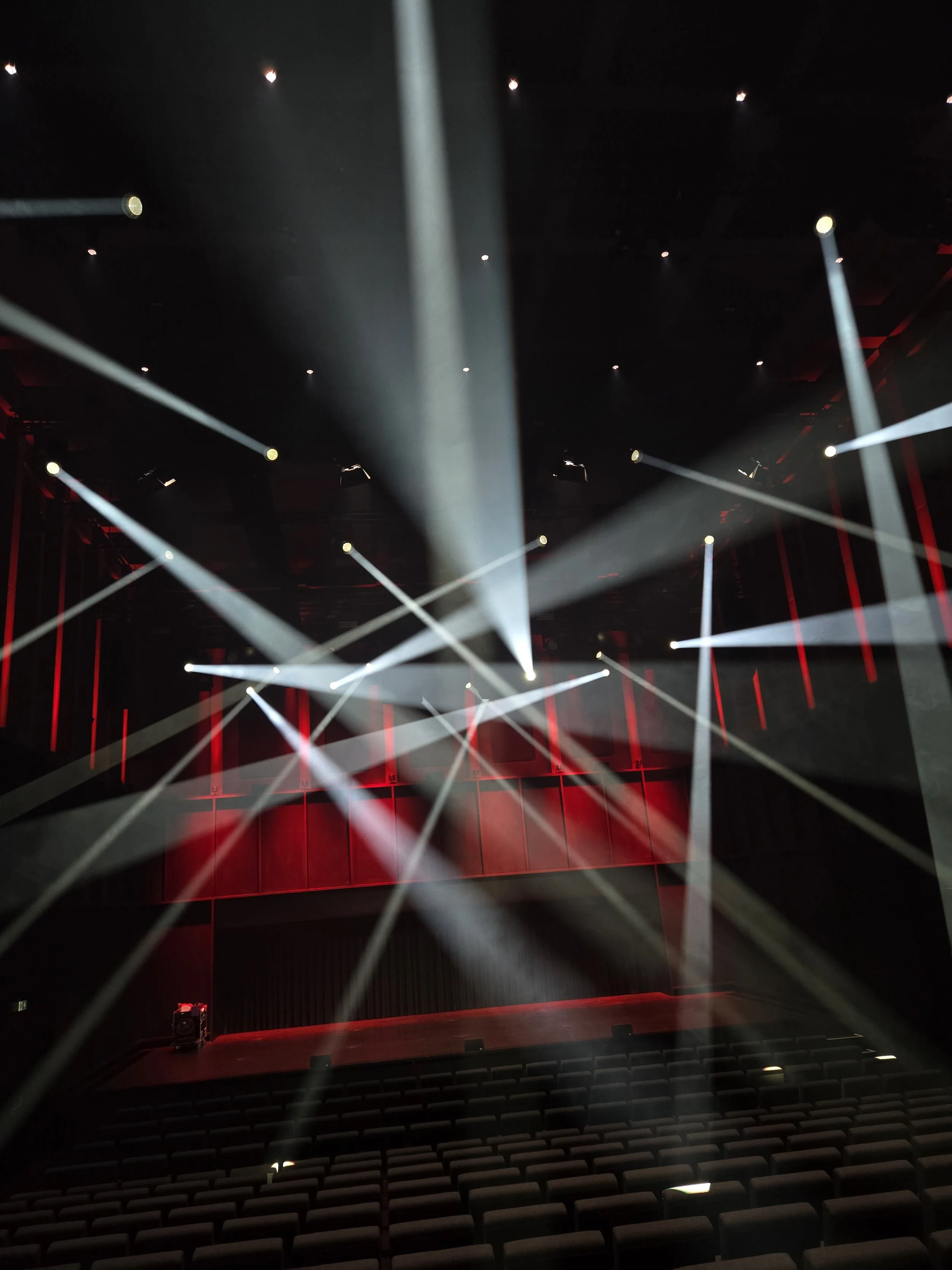 Empty auditorium or theater with rows of seats and stage, illuminated by stage lights and red lighting on the walls.