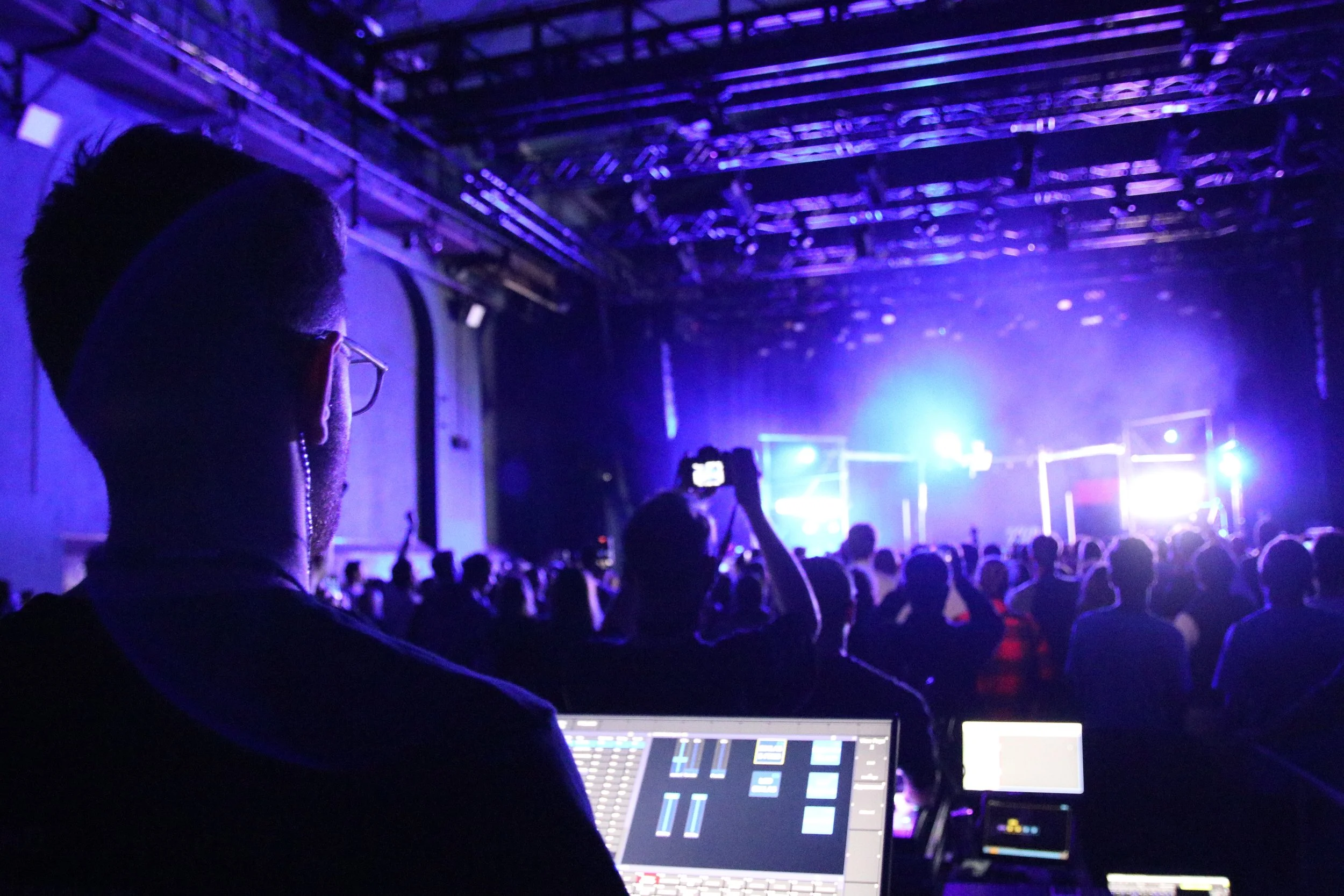 A person working with lighting controls at a concert, with a crowd and stage in the background illuminated by blue and purple lights.
