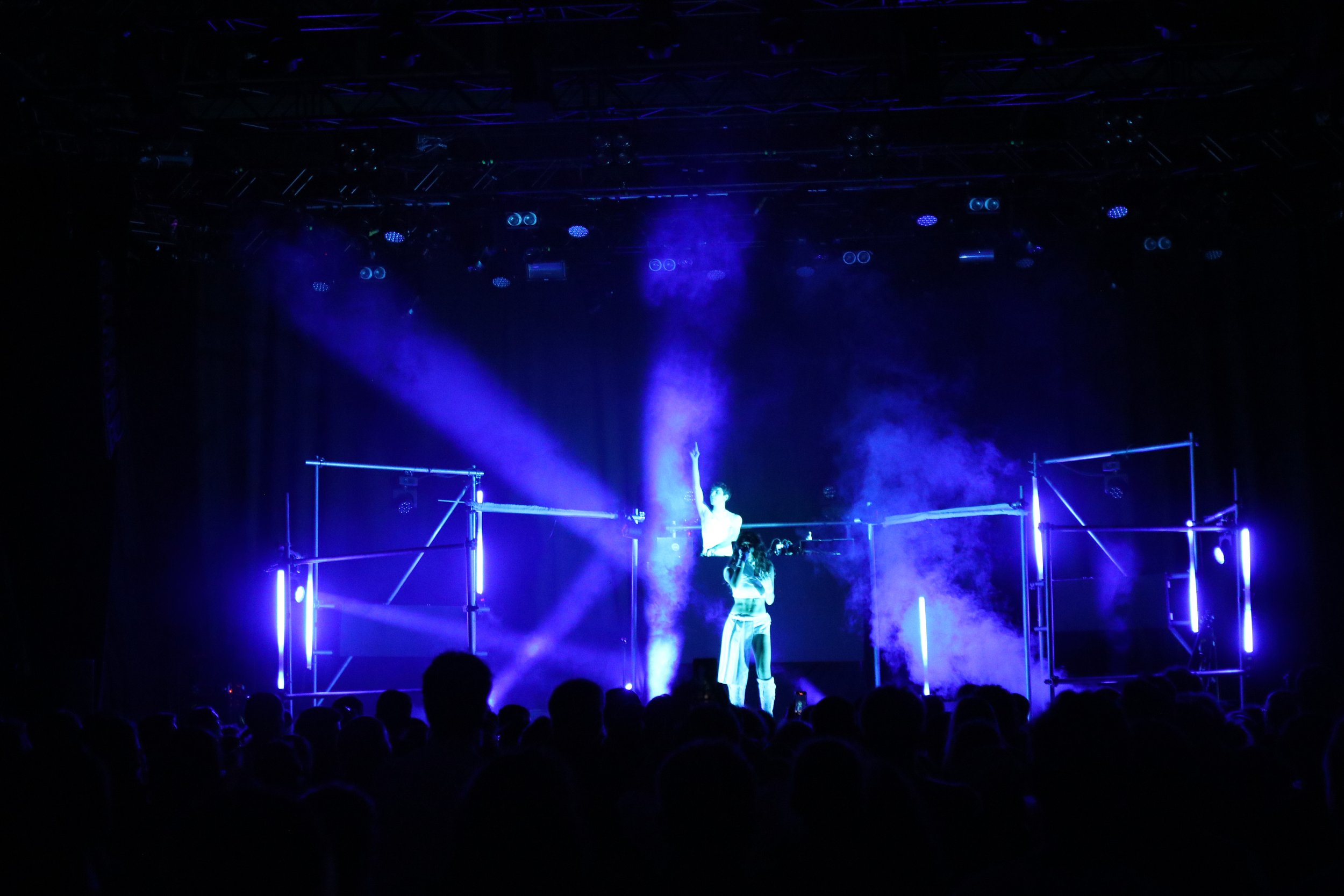 A dark stage with performers under blue and purple lighting, surrounded by metal scaffolding, in front of a large audience.