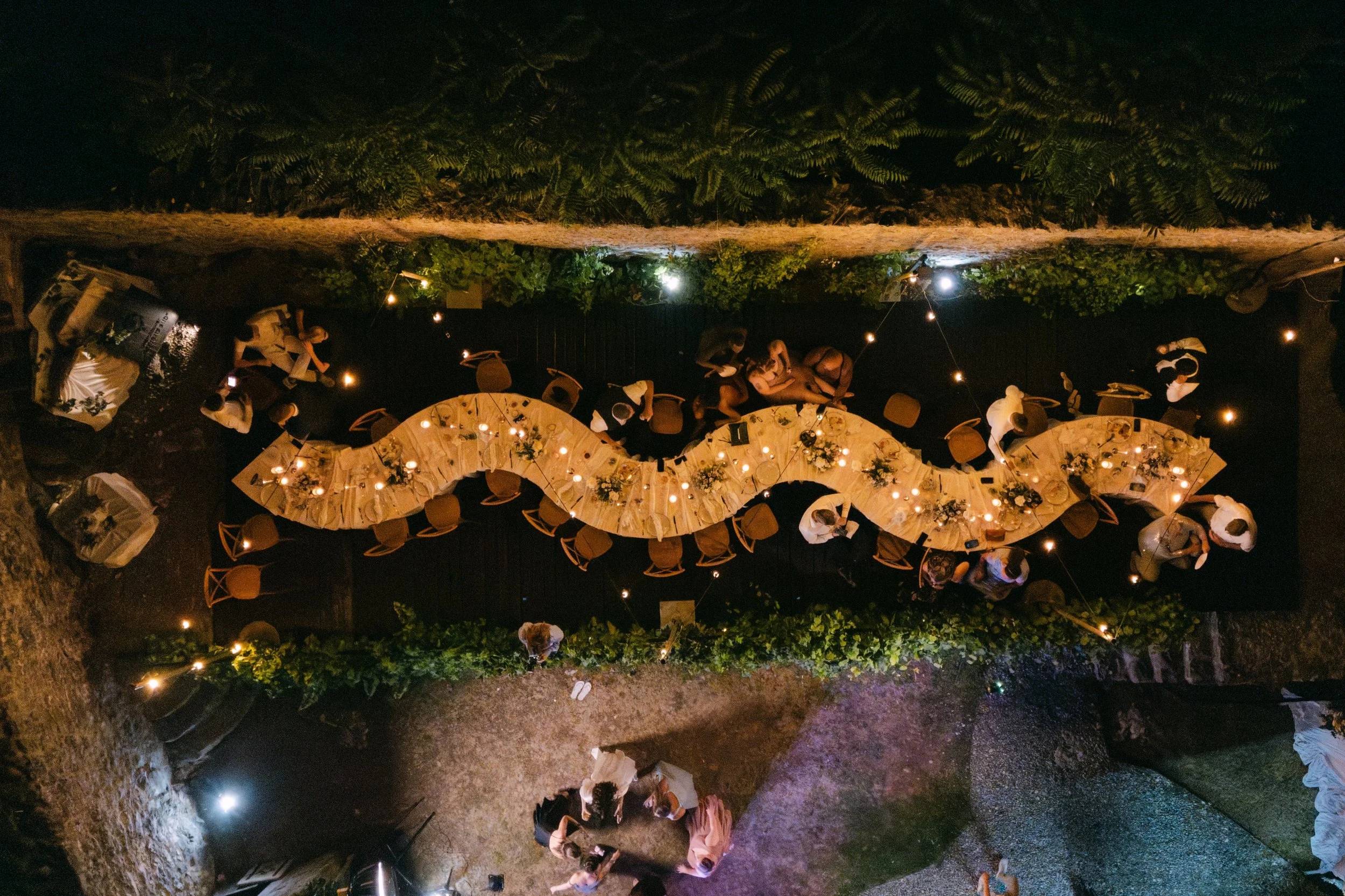 Night-time outdoor event with serpentine / snake tables decorated with flowers, surrounded by greenery and lighted by chandeliers