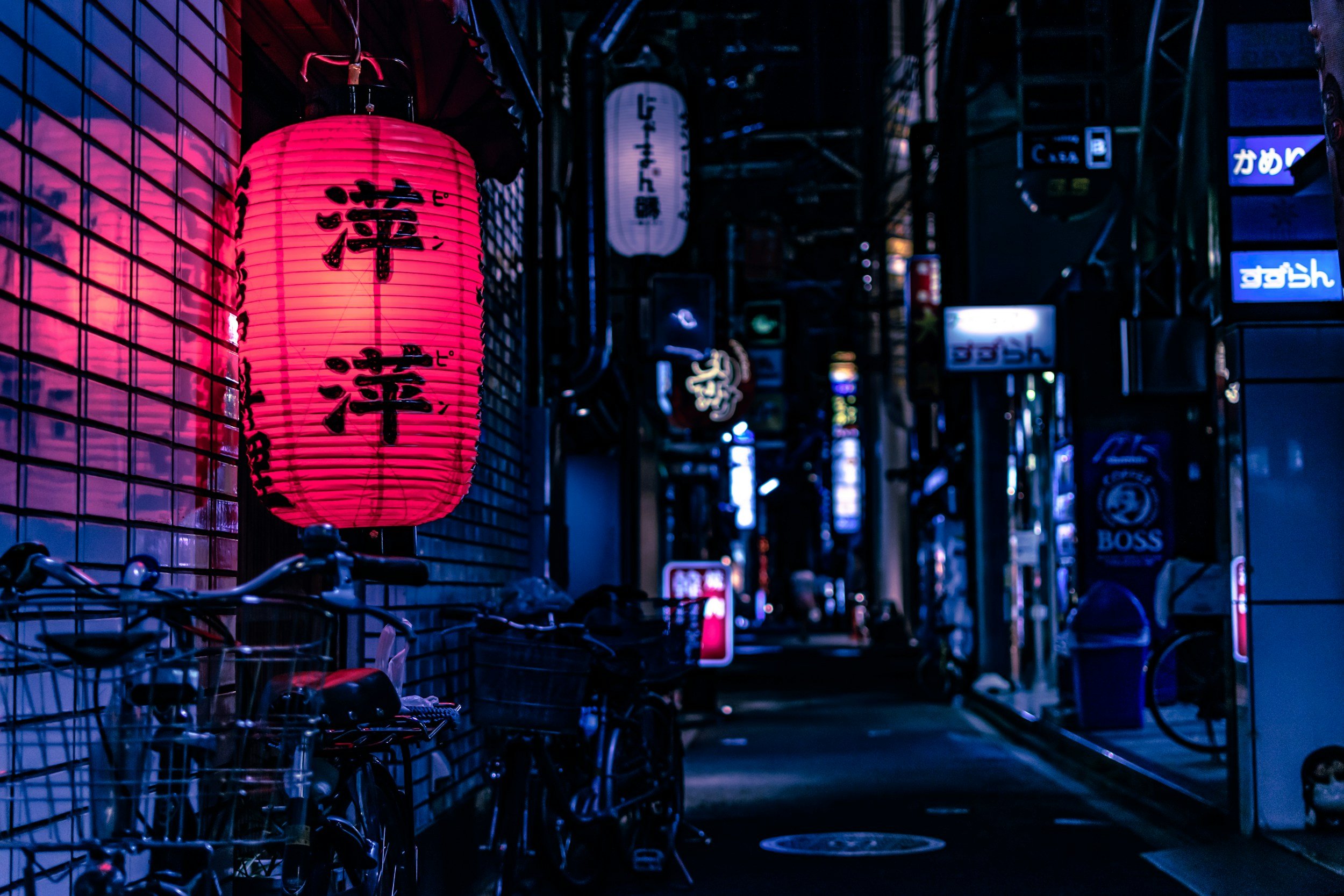 Night scene of a narrow alleyway illuminated by pink and blue neon lights, with Japanese paper lanterns hanging and bicycles parked along the sidewalk.