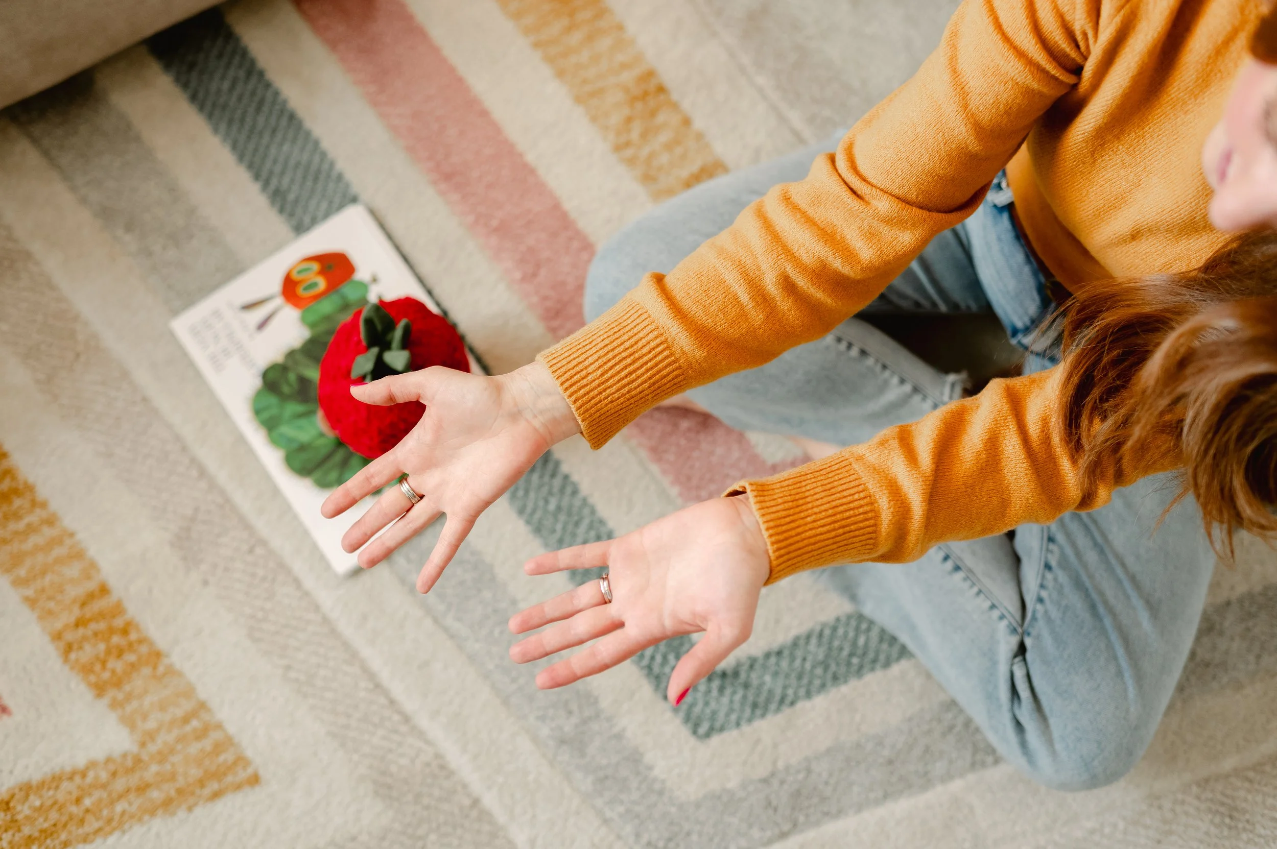 Person wearing an orange sweater and jeans sitting on a carpeted floor with a children's book with a caterpillar illustration and a plush toy resembling a red and green ladybug nearby.