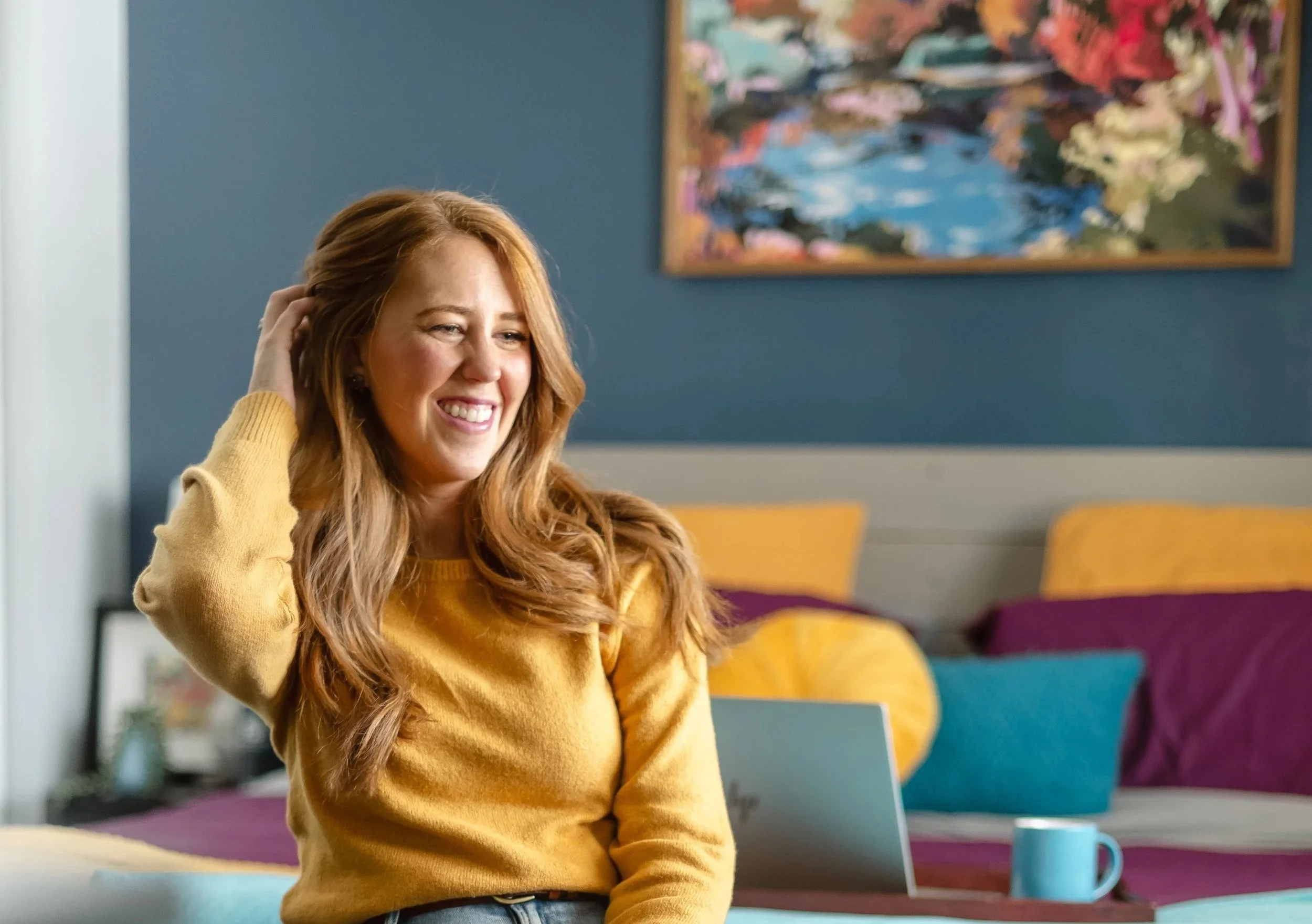 A woman with long red hair smiling while sitting on a bed with colorful pillows. She wears a yellow sweater and is holding her hair back with one hand. A laptop and a blue mug are on a tray in front of her. There is a framed painting on a blue wall in the background.