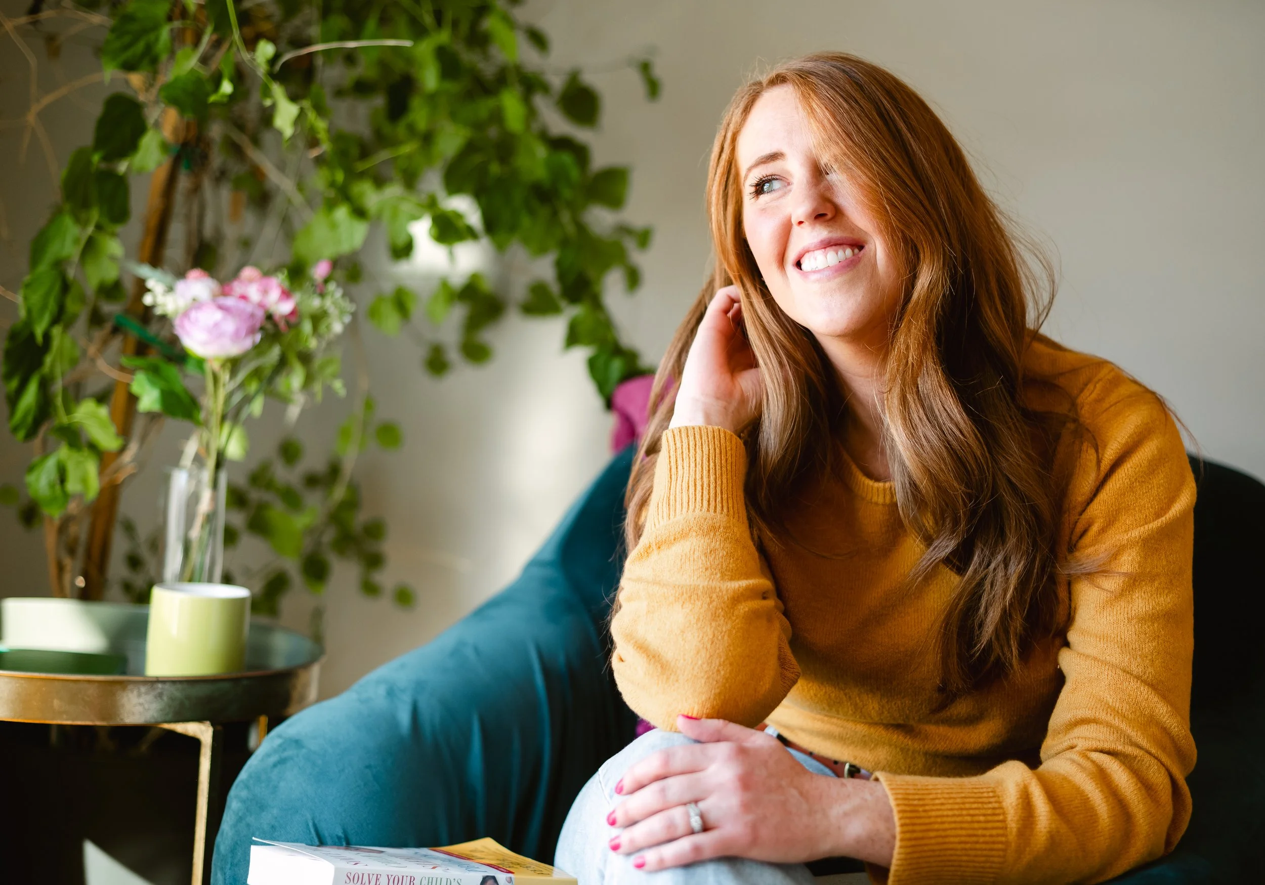 A smiling woman with long red hair sitting on a teal velvet couch next to a table with a magazine, in a room with a large green plant and flowers in a glass vase.