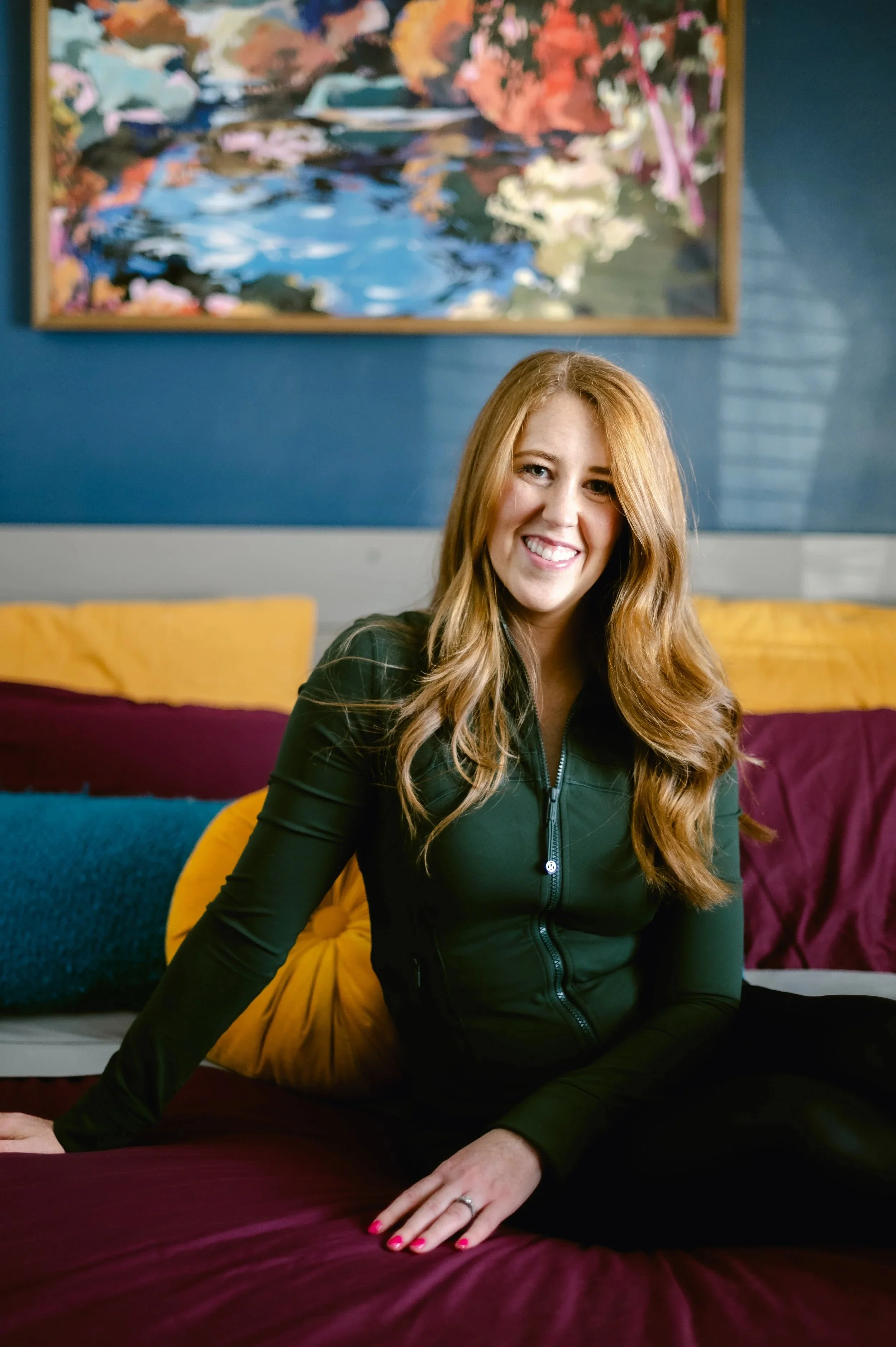 A young woman with long red hair, sitting on a bed with colorful pillows, smiling at the camera, in a room with a large abstract painting on a blue wall behind her.