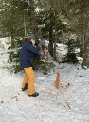 A person wearing a blue jacket, yellow pants, a gray beanie, and gloves pounding a wooden stake into snow-covered ground using a power tool in a forested area.