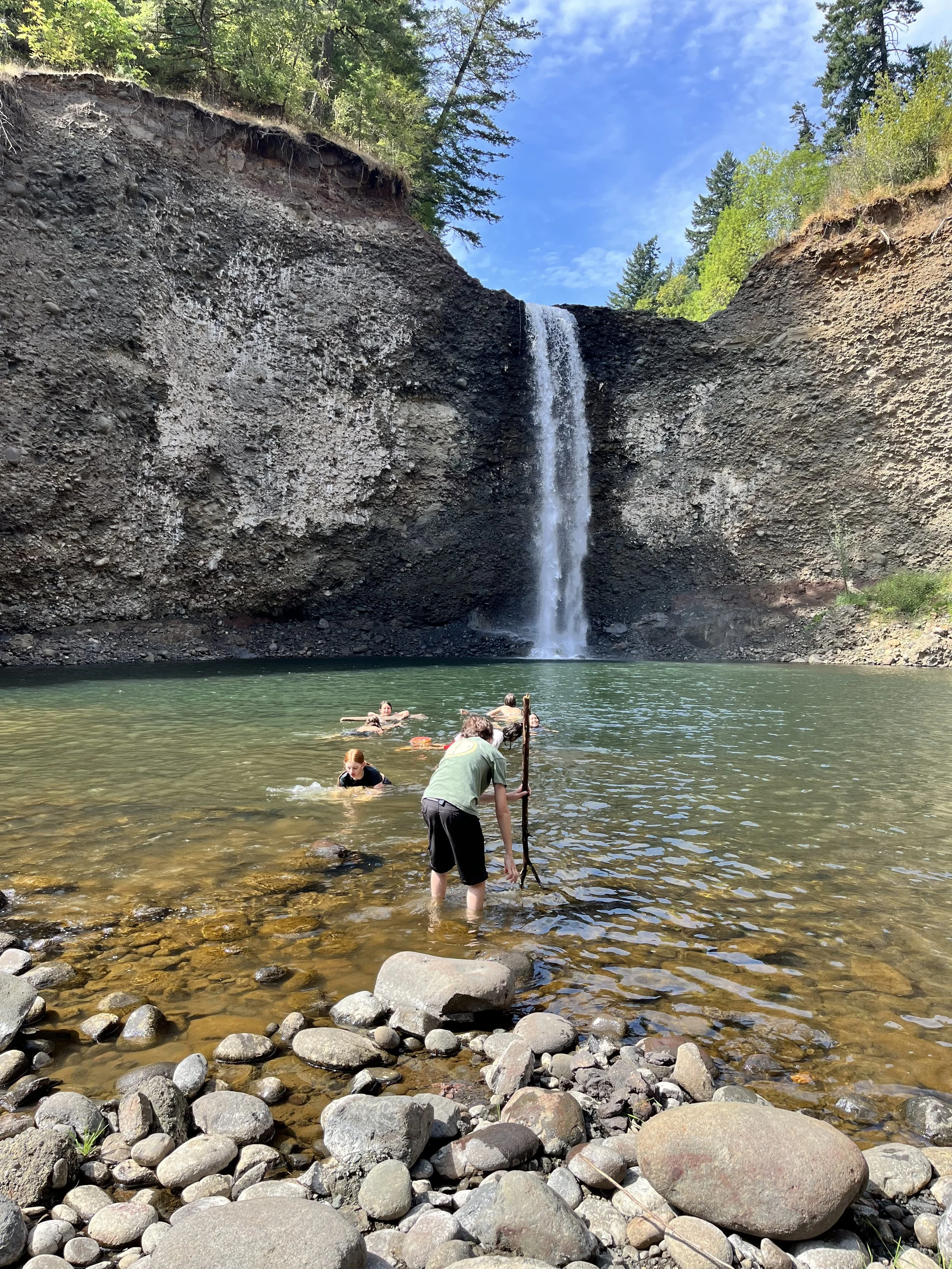 People swimming and standing in a river in front of a tall waterfall flowing down a rocky cliff surrounded by trees under a clear blue sky.