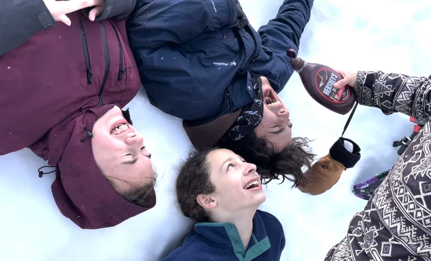 Four children lying on snow, smiling and laughing, with an adult pouring Hershey's chocolate syrup into their mouths.