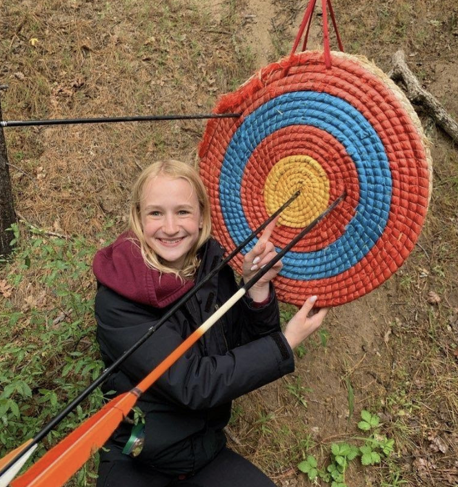 A smiling young girl in a black jacket and maroon hoodie poses in front of a colorful archery target on a wooded hillside, holding an arrow.