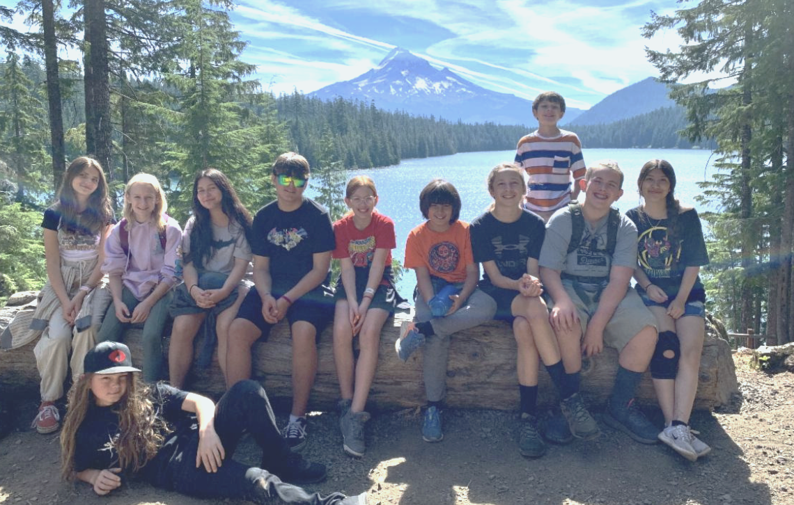 Group of kids sitting on a log by a lake with a snow-capped mountain and forest in the background.