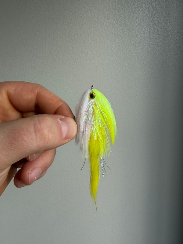 Hand holding a bright yellow and white fishing lure with feather and hair-like details against a plain light gray background.