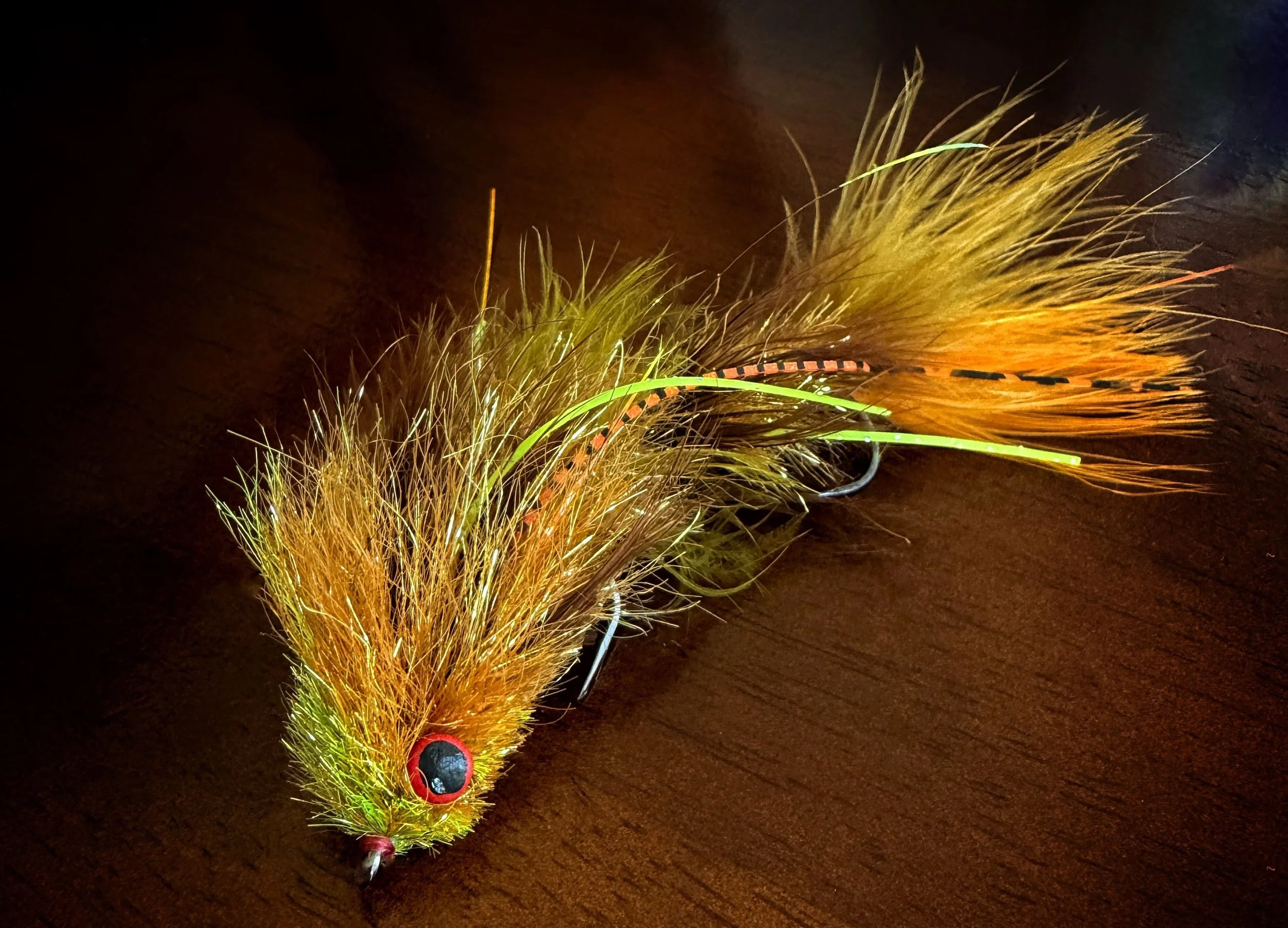 Close-up of a yellow and orange fishing lure with fur, feathers, and a red and black eye, lying on a wooden surface.