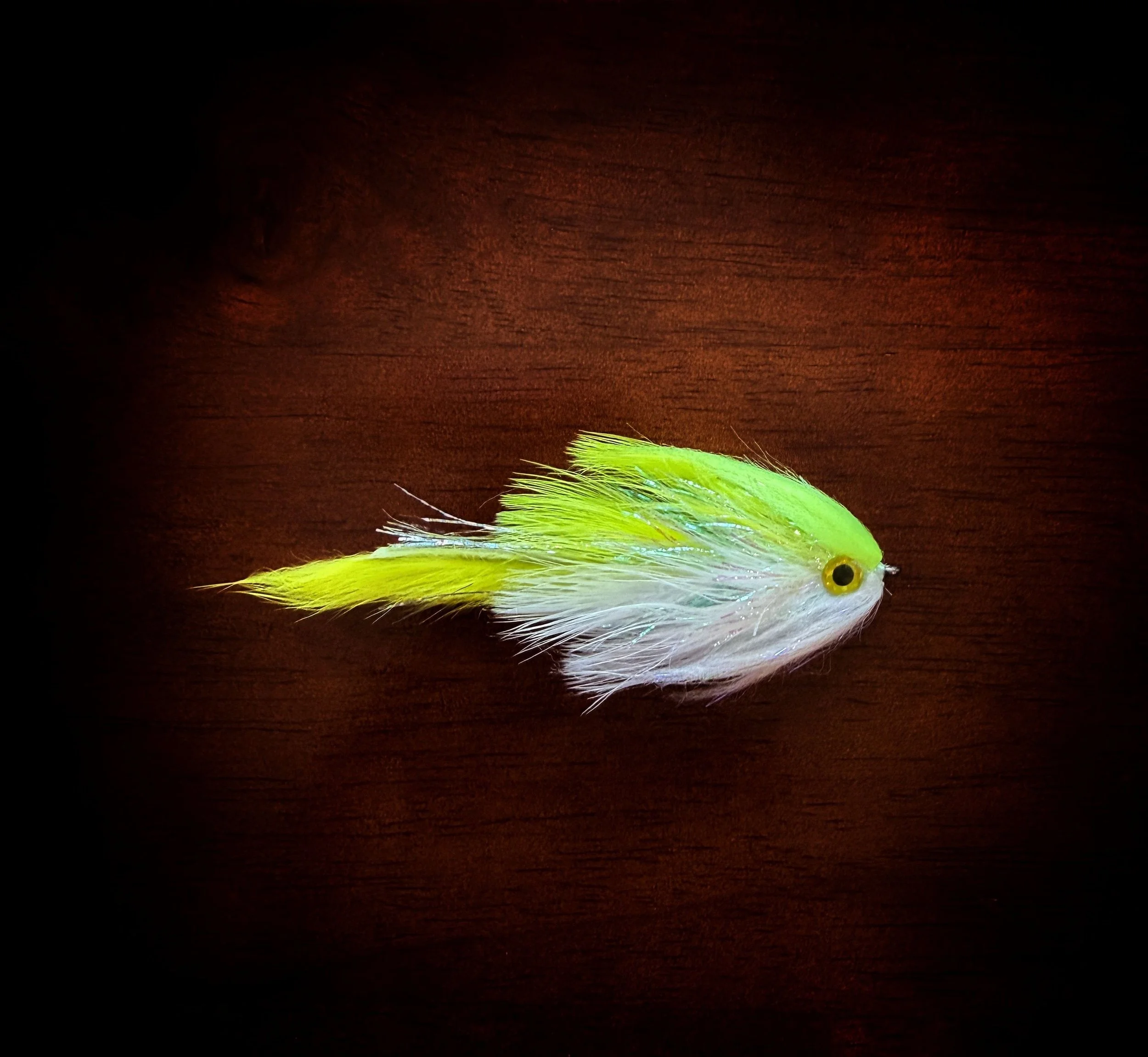 A fishing lure resembling a small fish, with bright green, yellow, and white feathers, lying on a dark wooden surface.